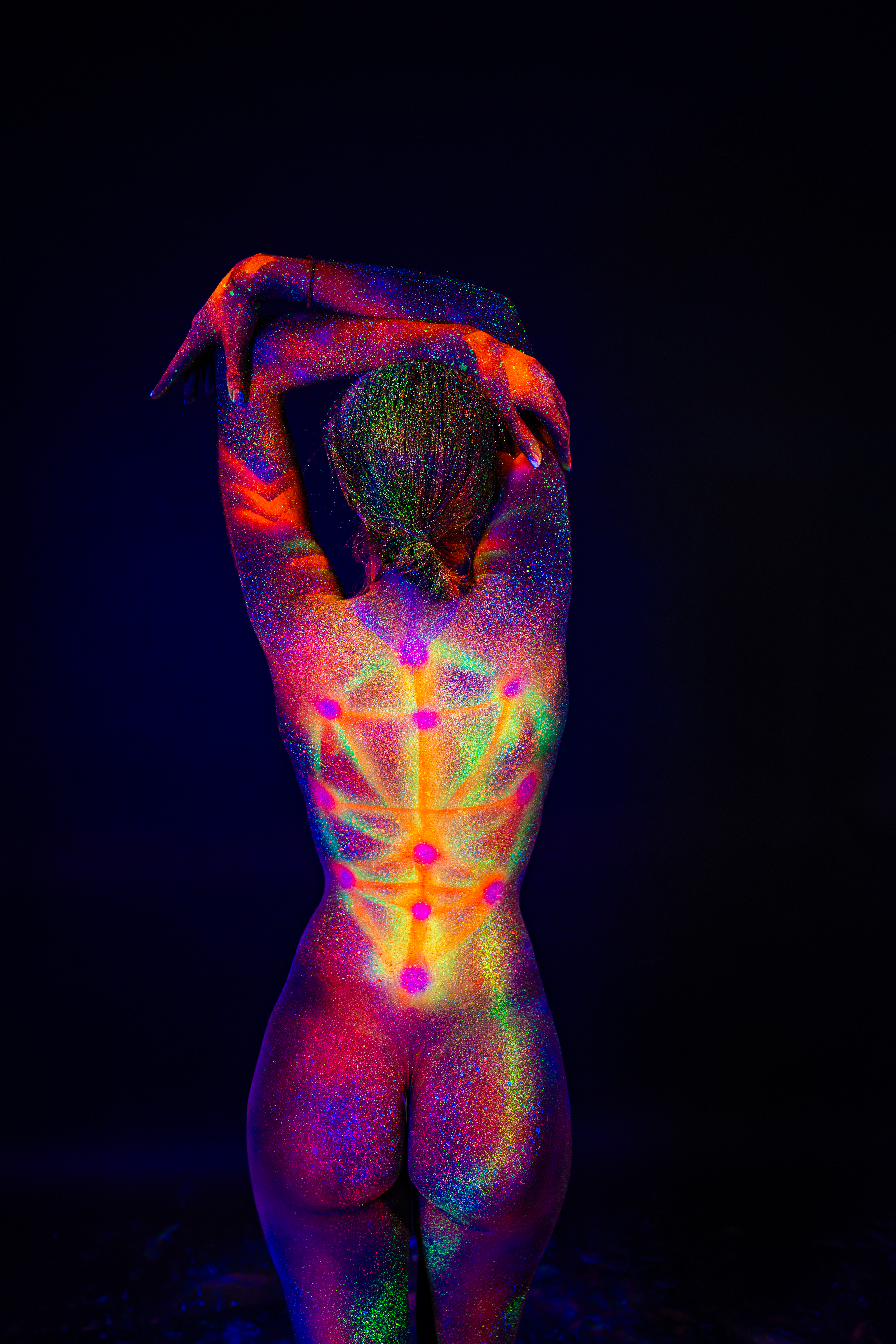 Sofia stands with her back to the camera in a full-length pose. A symmetrical geometric design of dots connected by radiating lines runs down her spine in glowing rainbow colors. Bodypaint by Elad Itzkin. Photographed by Elad Itzkin Photography under UV/blacklight in his studio in Gesundbrunnen, Berlin.