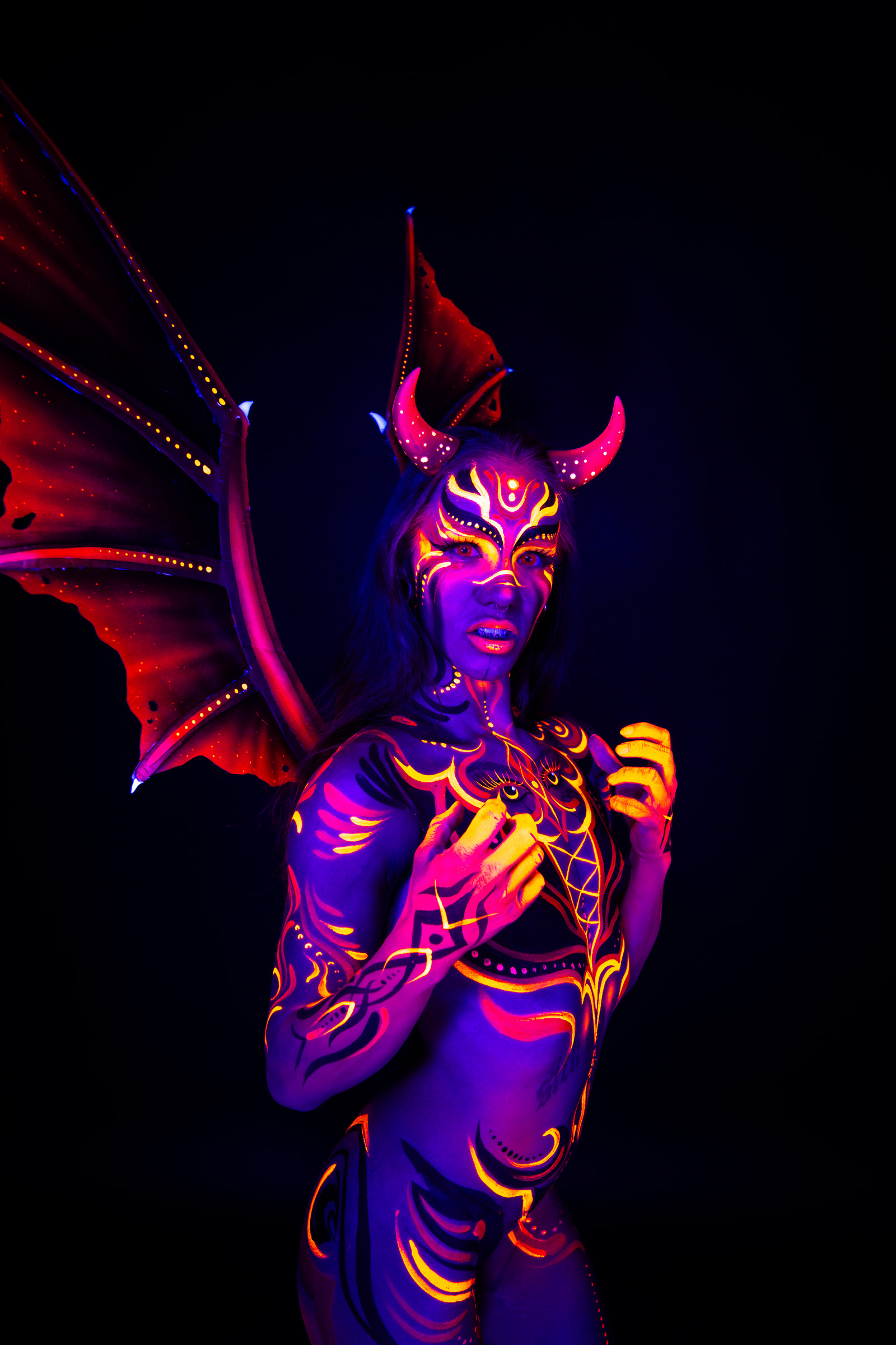 Thory stands in a three-quarter pose with wing shaped headpiece elements spread behind the shoulders. The bodypaint is a devil themed design in red, orange and purple tones with horn motifs and geometric linework. Bodypaint by Joana Rohr. Photographed by Elad Itzkin Photography under UV light in his studio in Gesundbrunnen, Berlin.