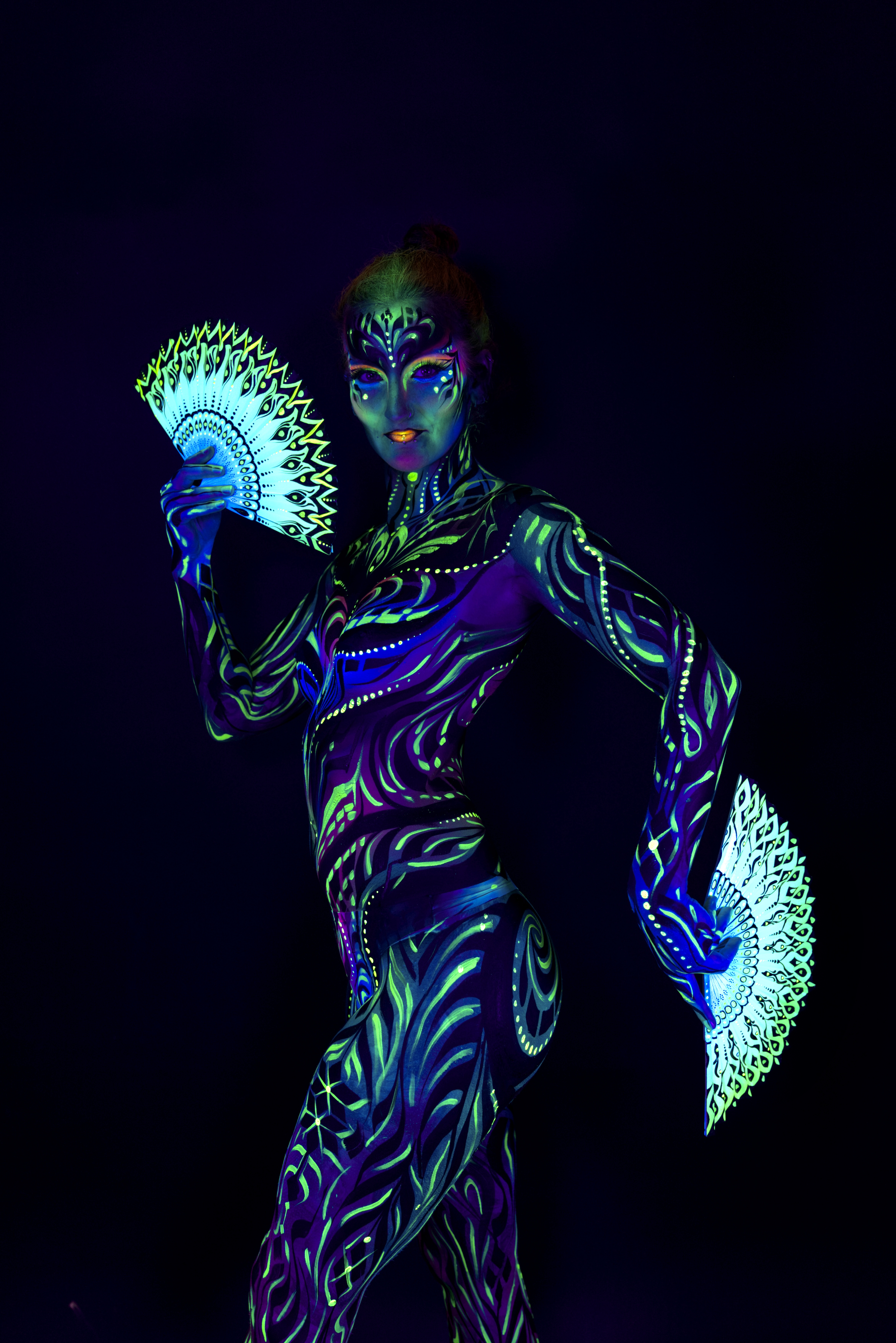 Joey stands in a three-quarter pose facing the camera, holding two fan-shaped props. The bodypaint is a blue and green swirling linework pattern covering the torso and limbs. Bodypaint by Joana Rohr and Zami. Photographed by Elad Itzkin Photography under UV light in his studio in Gesundbrunnen, Berlin.