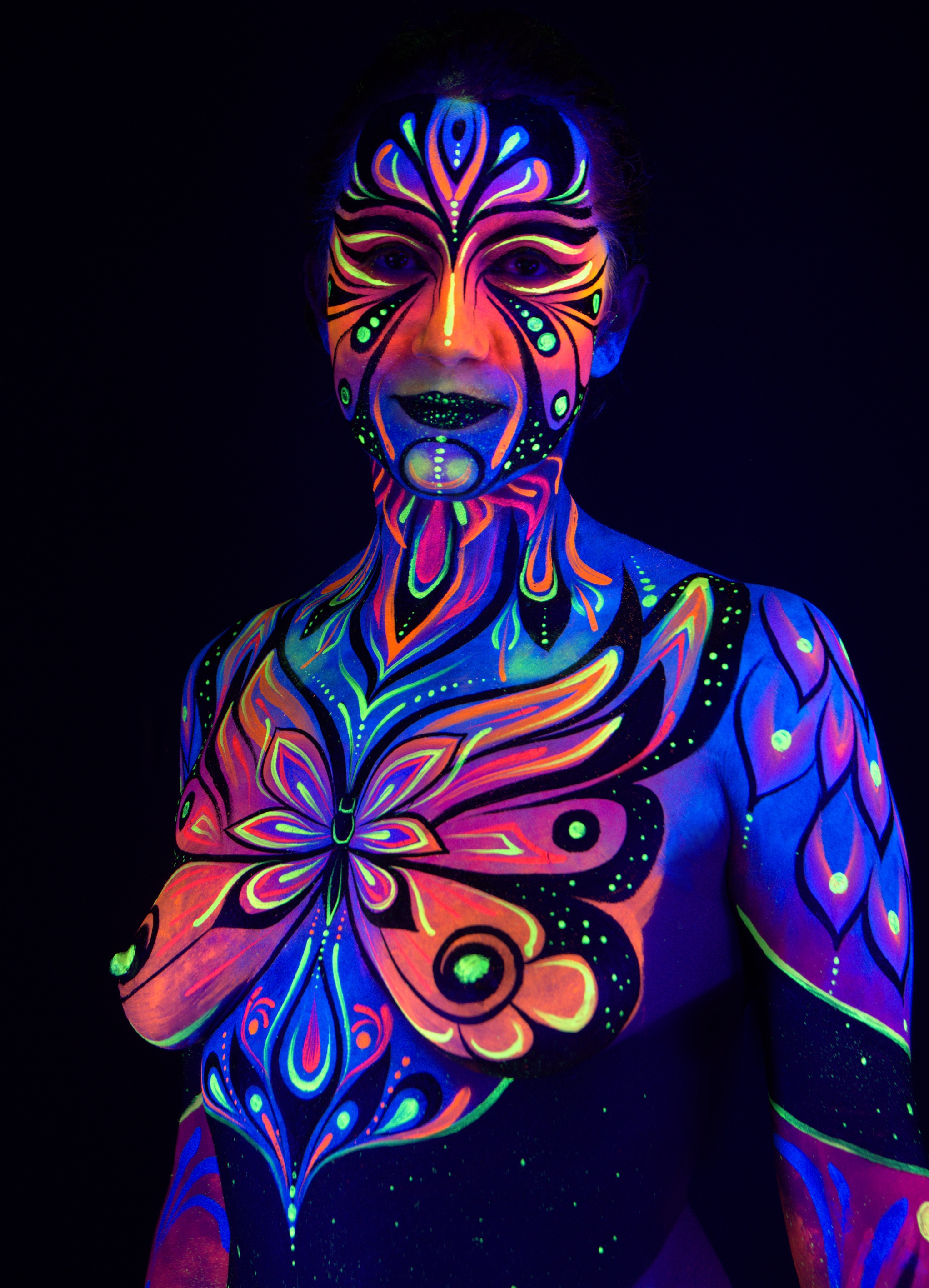 Katrina stands facing the camera in an upper-body crop. A butterfly-shaped design in pink, orange, and blue covers her chest, extending into matching linework on the face and shoulders. Bodypaint by Joana Rohr. Photographed by Elad Itzkin Photography under UV light in his studio in Gesundbrunnen, Berlin.