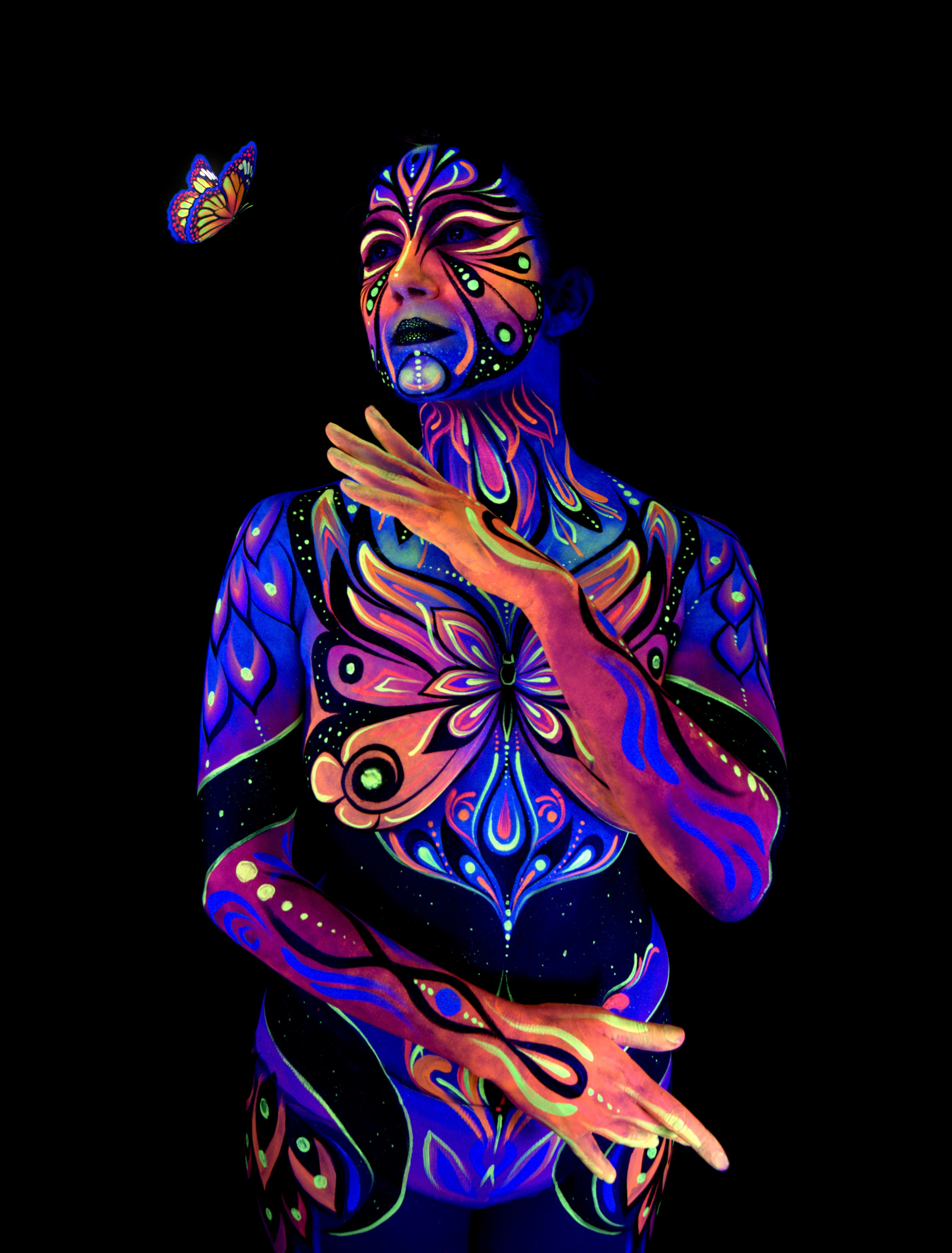 Katrina stands in an upright pose, facing slightly upward. Her chest is painted with a butterfly-shaped design in pink, orange, and blue, matched by coordinating linework on the face and shoulders. Bodypaint by Joana Rohr. Photographed by Elad Itzkin Photography under UV light in his studio in Gesundbrunnen, Berlin.