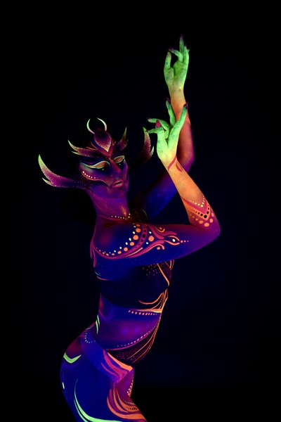 Rala du Reign stands in a three-quarter pose, body angled toward the camera. The design is a purple and orange tribal-style pattern with a horned, mask-like face and abstract dotted linework across the torso. Bodypaint by Joana Rohr. Photographed by Elad Itzkin Photography under UV light in his studio in Gesundbrunnen, Berlin.