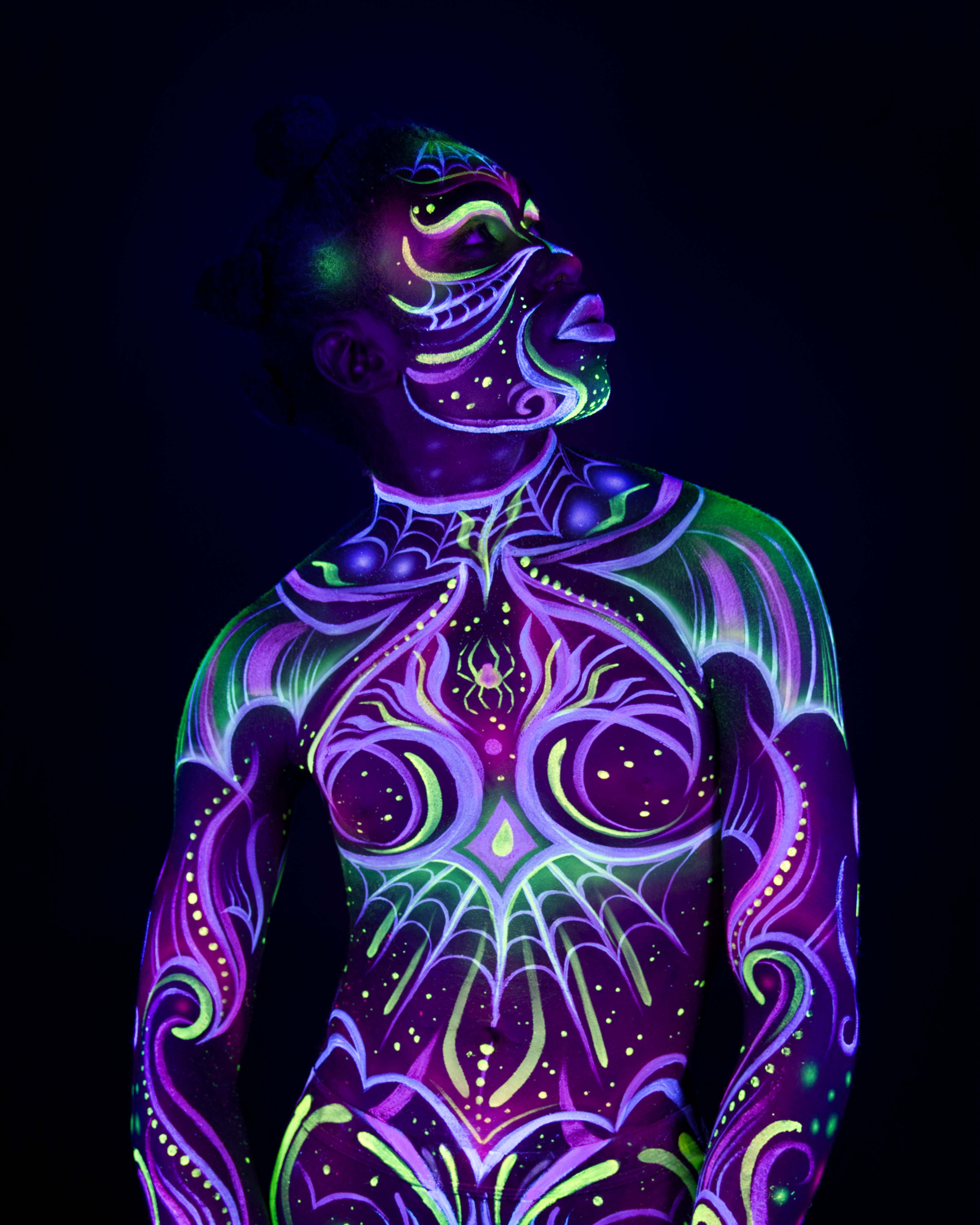Bikme stands with her head tilted back, face turned to the side. The bodypaint design covers her face, neck, and chest in a purple and green web-like pattern, including a small spider motif at the center of the chest. Bodypaint by Joana Rohr and Sage Brewer. Photographed by Elad Itzkin Photography under UV light in his studio in Gesundbrunnen, Berlin.