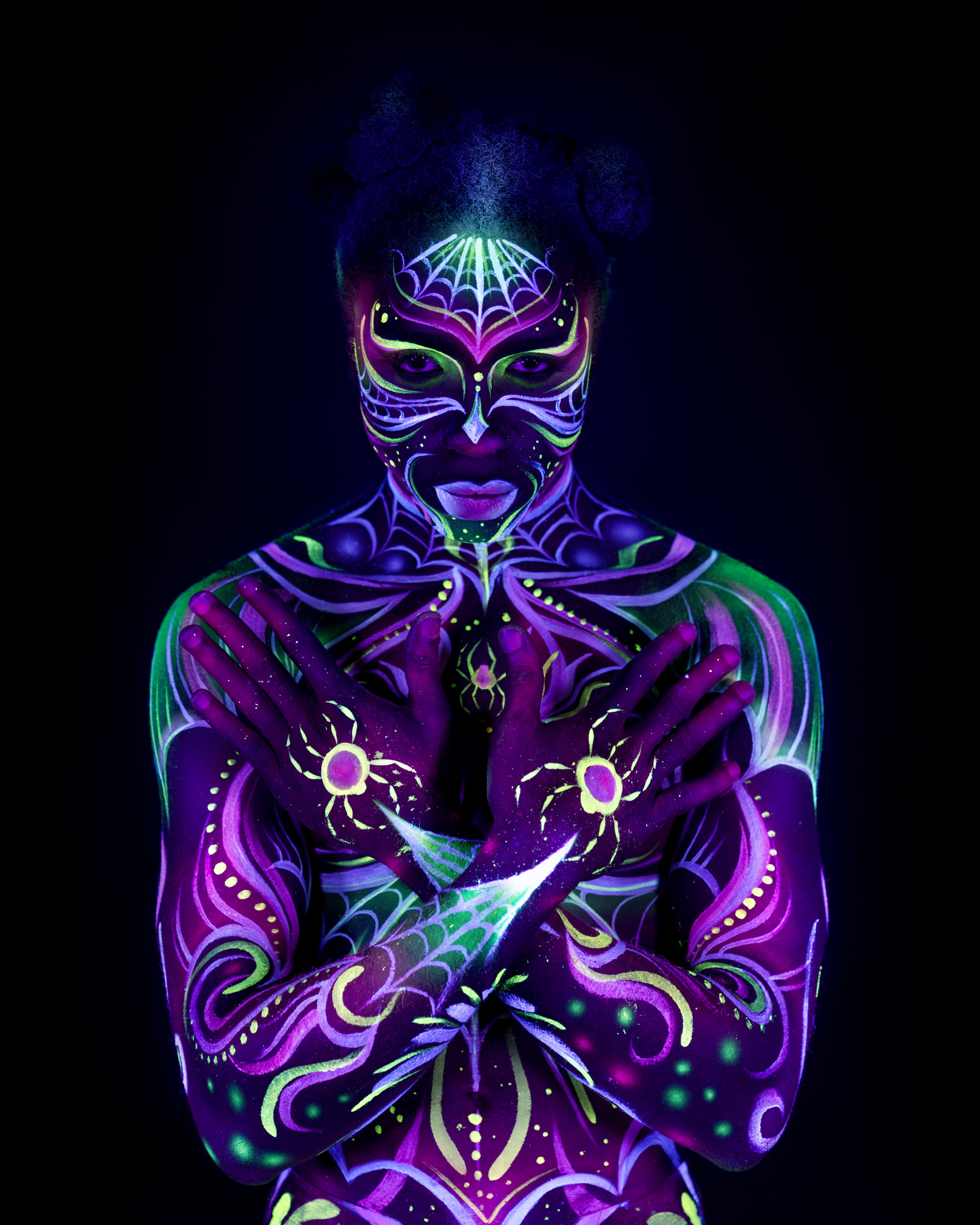 Bikme stands facing the camera in an upright pose. The design is a purple and green web-like pattern across the face, neck, and chest, featuring a spider motif and matching mask-like facial linework. Bodypaint by Joana Rohr and Sage Brewer. Photographed by Elad Itzkin Photography under UV light in his studio in Gesundbrunnen, Berlin.