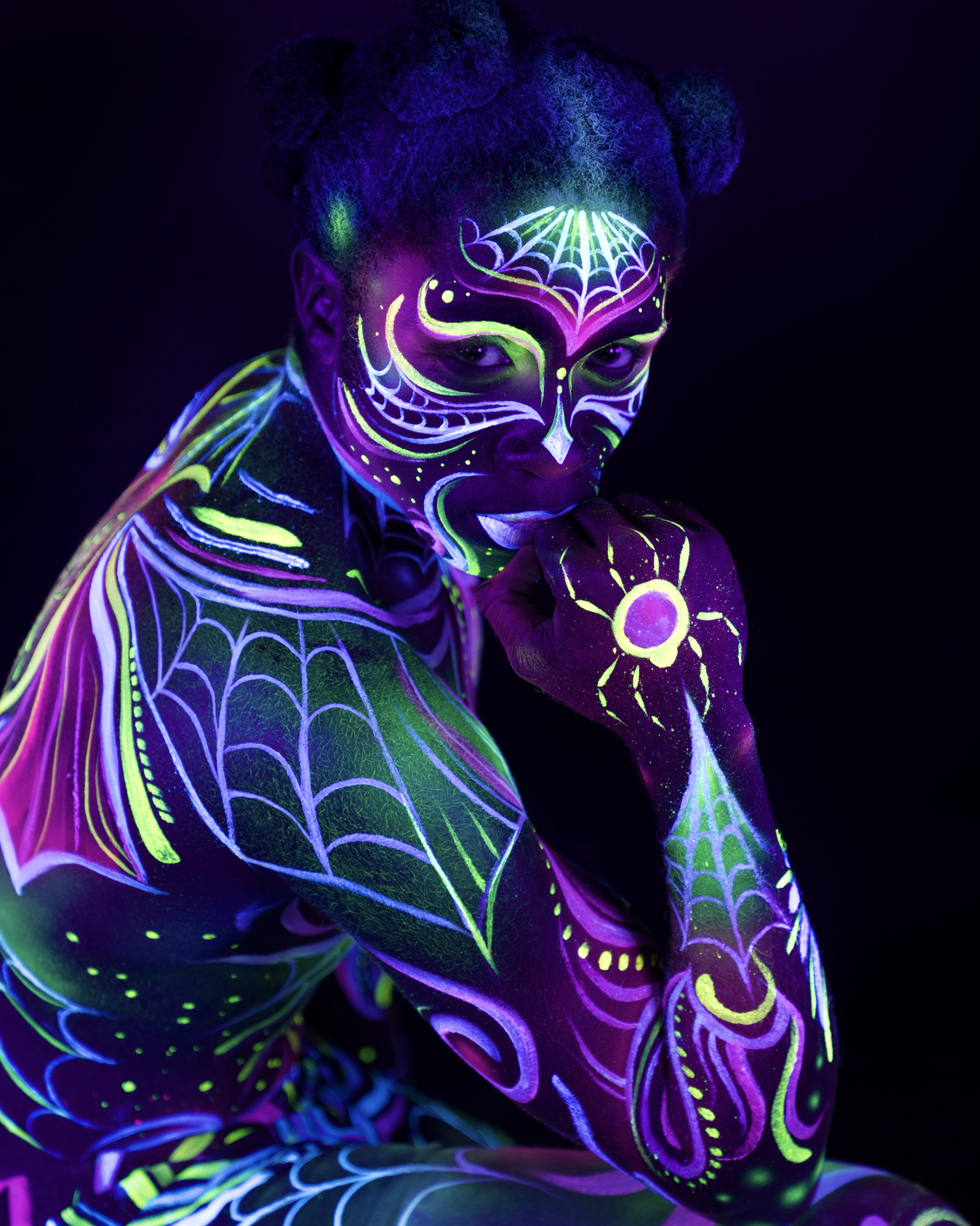 Bikme is shown in a close, three-quarter pose from the waist up, body turned toward the camera in a crouched position. Purple and green spiderweb and floral linework covers the face and shoulder, with a small sun motif on the chest. Bodypaint by Joana Rohr, Sage Brewer. Photographed by Elad Itzkin Photography under UV light in his Gesundbrunnen, Berlin studio.