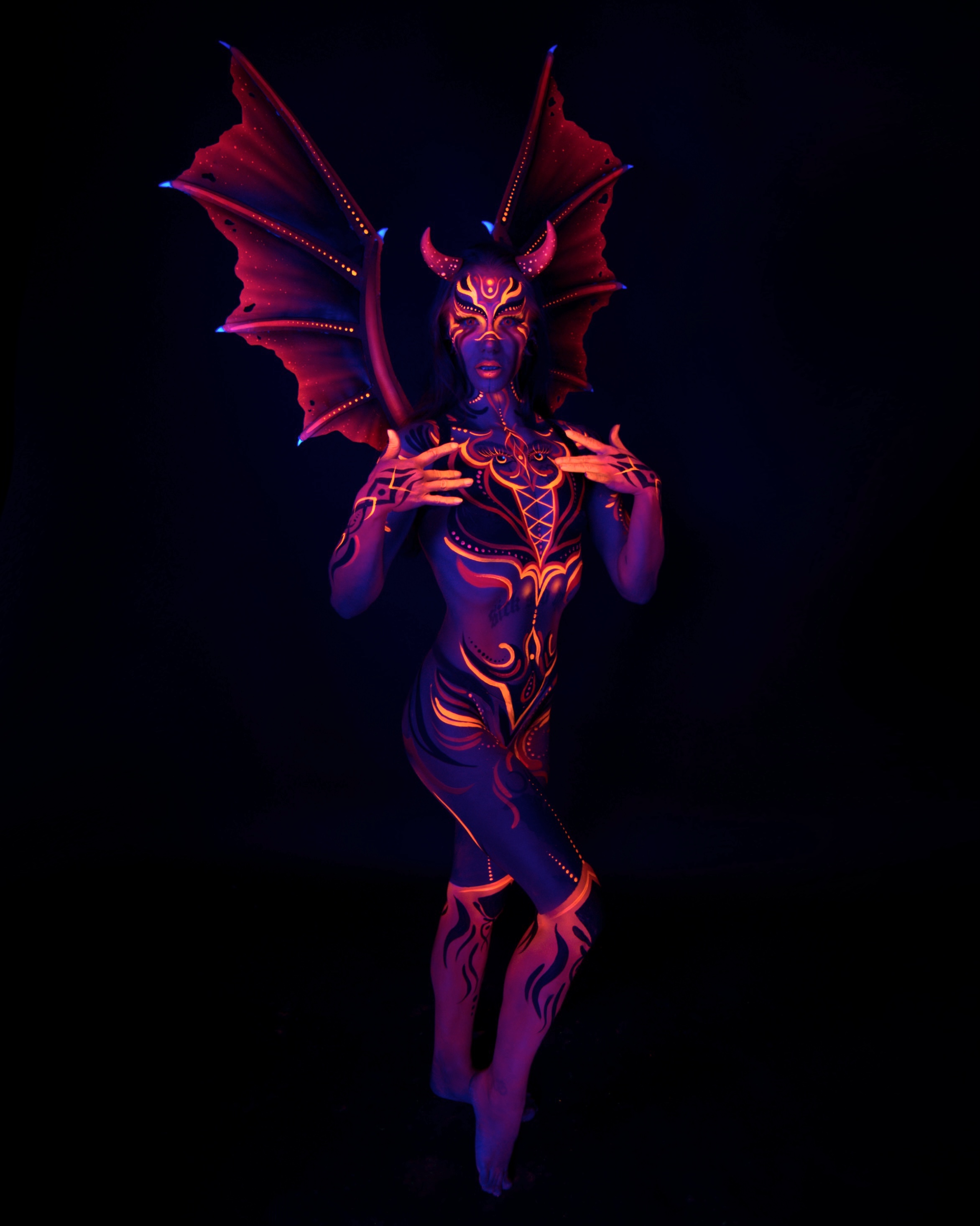 Thory stands facing the camera in a full length frontal pose. The design features large red and orange bat-like wings and horns on the head, with orange and purple geometric linework extending down the body. Bodypaint by Joana Rohr. Photographed by Elad Itzkin Photography under UV light in his Gesundbrunnen, Berlin studio.
