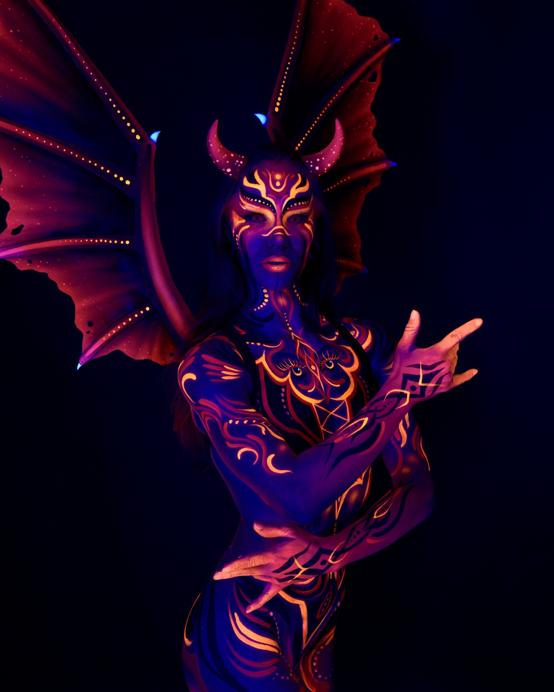 Thory is photographed standing and facing forward, upper body visible, wearing a horned and winged headpiece. An orange and purple geometric design with eye-like detailing covers the chest and face. Bodypaint by Joana Rohr. Photographed by Elad Itzkin Photography under UV light in his Gesundbrunnen, Berlin studio.