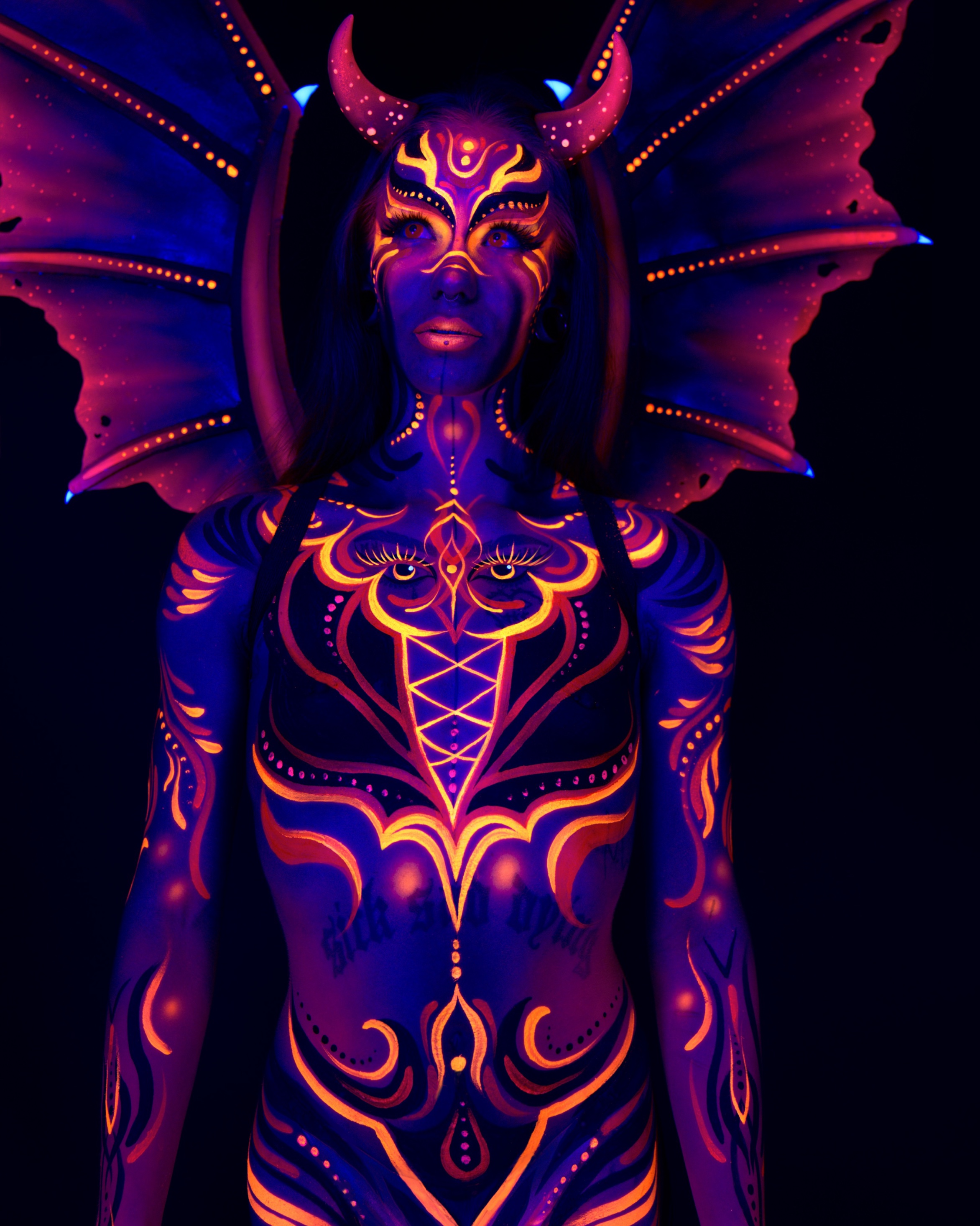 Thory stands facing the camera in a waist-up pose, wearing a horned headpiece with wings spread behind the shoulders. Orange and purple linework in a symmetrical pattern covers the chest and face. Bodypaint by Joana Rohr. Photographed by Elad Itzkin Photography under UV light in his Gesundbrunnen, Berlin studio.