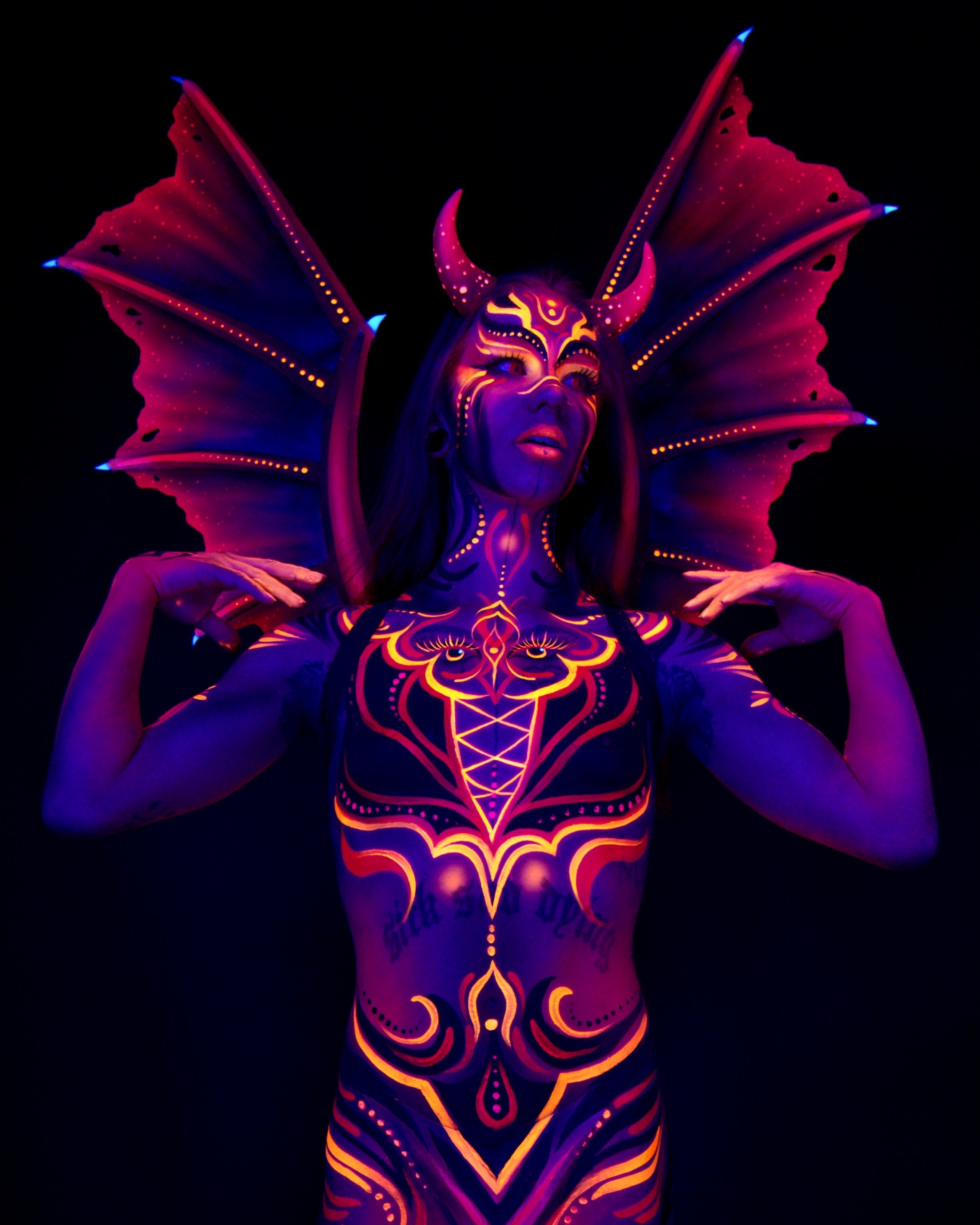 Thory faces the camera in an upper body pose with wings spread wide behind the shoulders and a horned headpiece. The chest and face carry a symmetrical orange and purple pattern with an eye-like design at center. Bodypaint by Joana Rohr. Photographed by Elad Itzkin Photography under UV light in his Gesundbrunnen, Berlin studio.