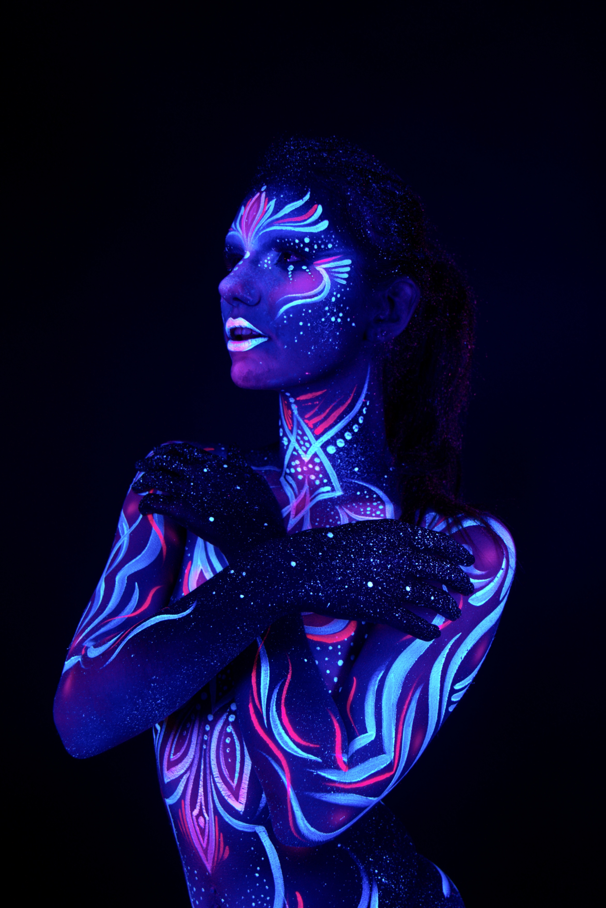 Nina stands with her body turned to the side in a three-quarter pose, facing away from the camera. Blue, pink and white linework in a floral pattern covers the torso and lower body. Bodypaint by Joana Rohr. Photographed by Elad Itzkin Photography under UV light in his Gesundbrunnen, Berlin studio.