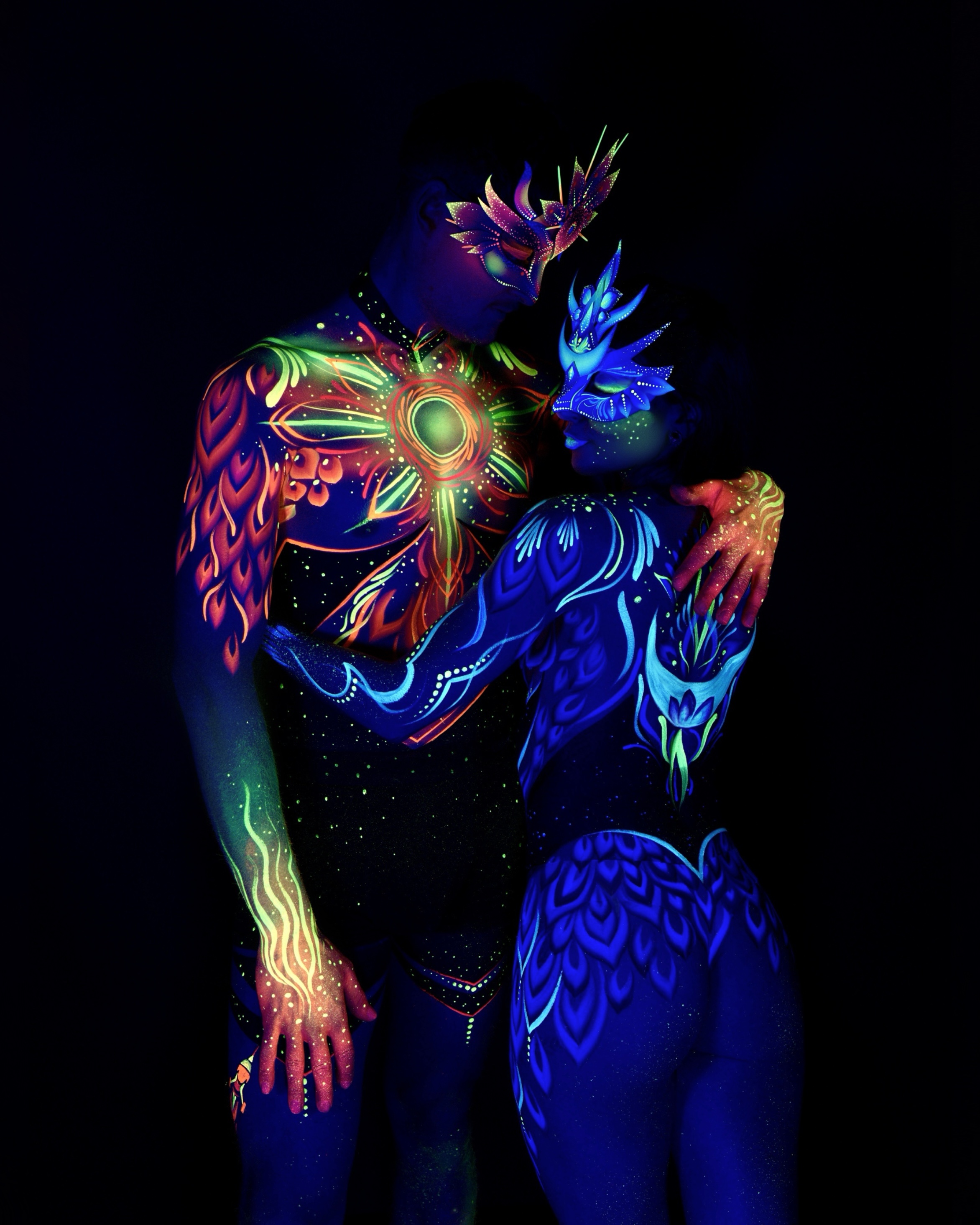 Sori and Julius stand close together in an embrace, Julius facing the camera and Sori turned toward him in profile. Julius wears an orange and green sunburst design on the chest, while Sori's face carries a blue crown-shaped headpiece with matching feather linework. Bodypaint by Joana Rohr. Photographed by Elad Itzkin Photography under UV light in his Gesundbrunnen, Berlin studio.