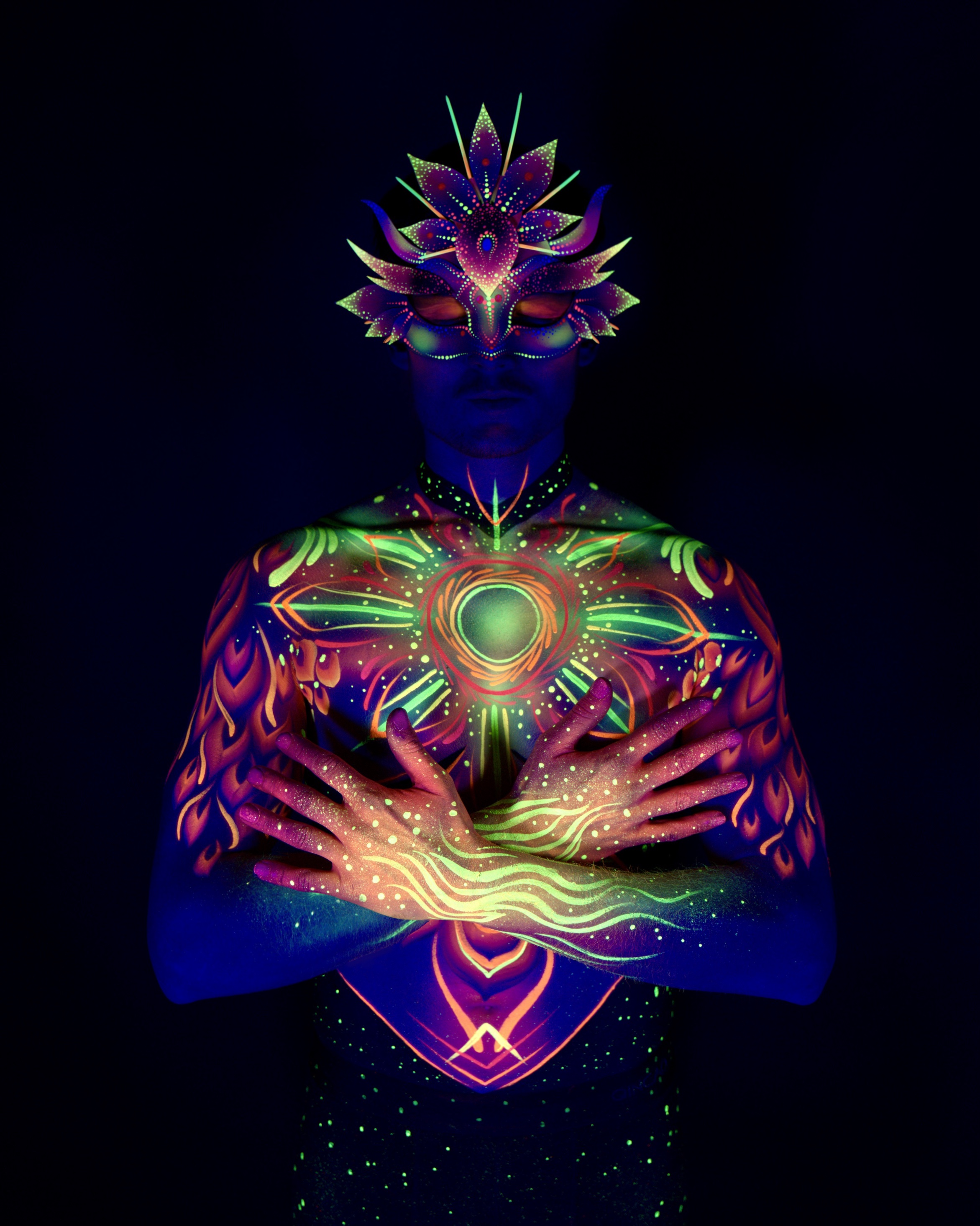 Julius stands facing the camera in a close upper body pose, wearing a spiked crown-shaped headpiece. The chest is painted with a green and orange sunburst and mandala design. Bodypaint by Joana Rohr. Photographed by Elad Itzkin Photography under UV light in his Gesundbrunnen, Berlin studio.