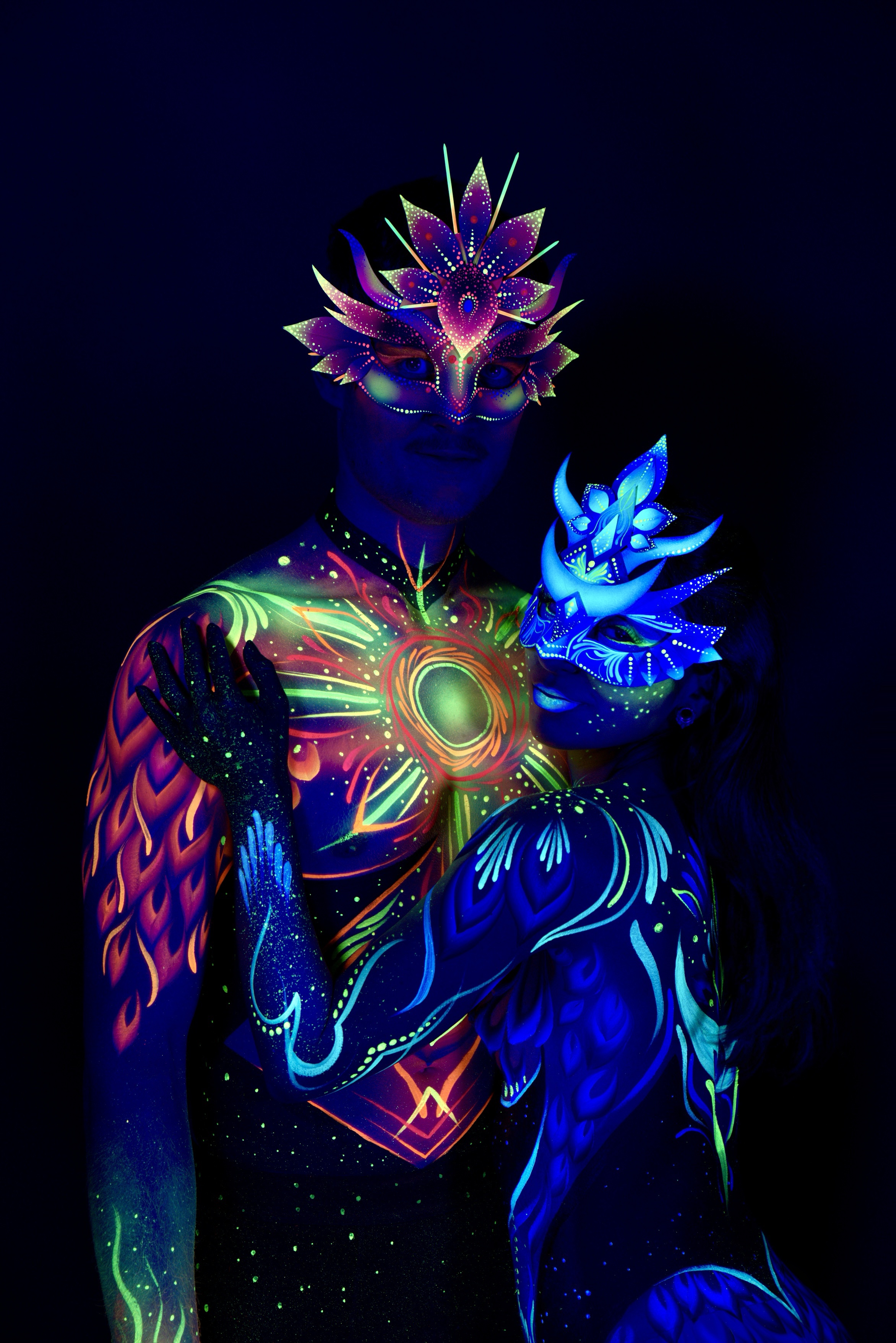 Sori and Julius stand close together in profile, Julius facing forward with an orange and green sunburst design across the chest. Sori stands beside him wearing a blue crown-shaped headpiece with matching blue linework. Bodypaint by Joana Rohr. Photographed by Elad Itzkin Photography under UV light in his Gesundbrunnen, Berlin studio.