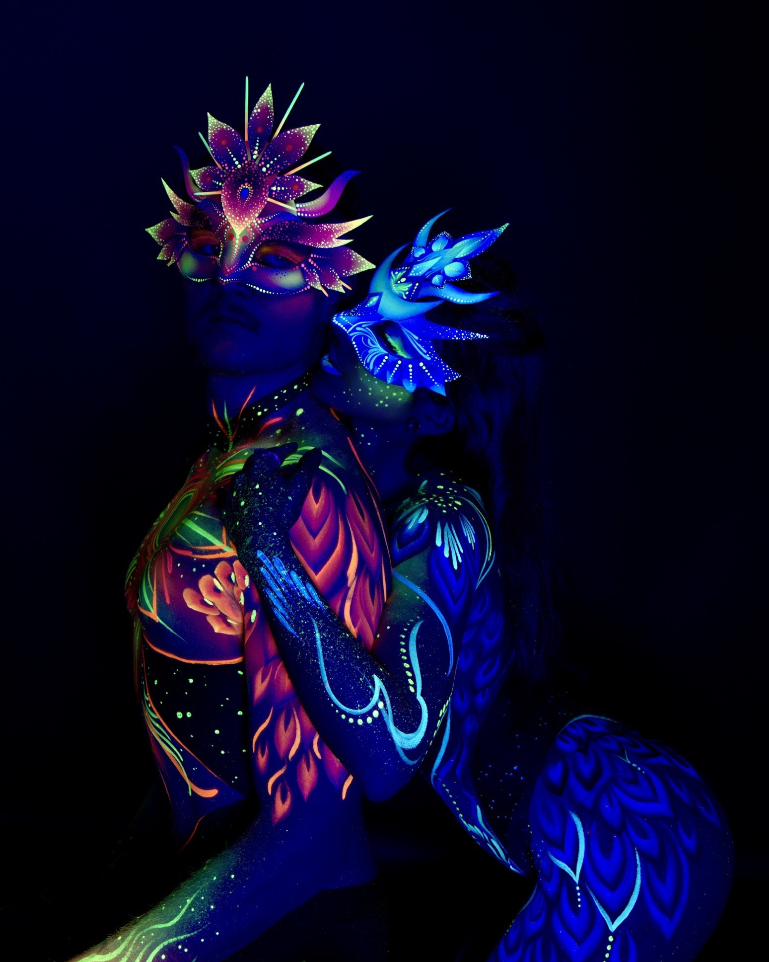 Sori and Julius stand close together in profile, with their backs partly turned to the camera. Julius's shoulder carries an orange and green floral pattern, while Sori's side and hip show a blue feather-like design. Bodypaint by Joana Rohr. Photographed by Elad Itzkin Photography under UV light in his Gesundbrunnen, Berlin studio.