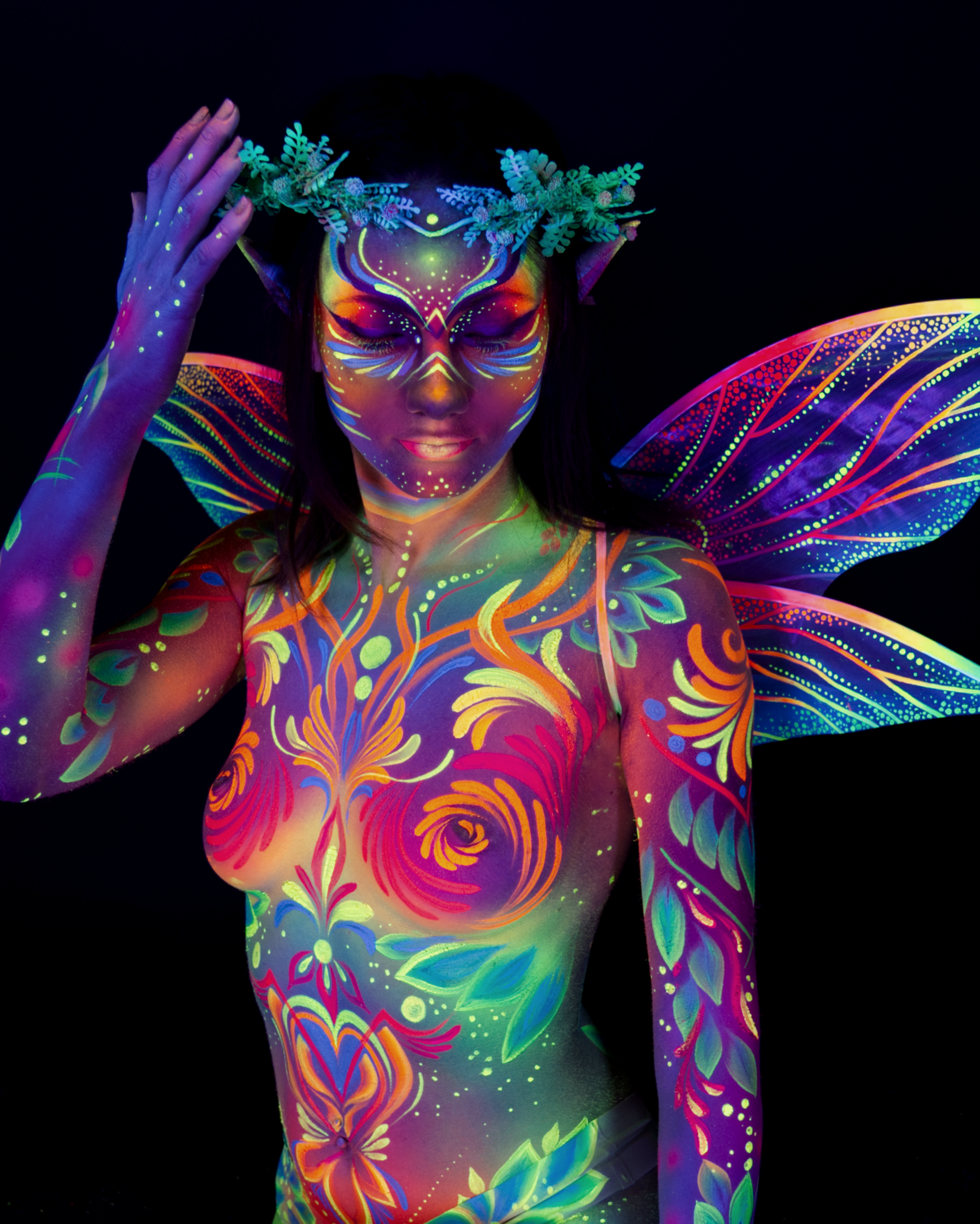 Marina stands in a three-quarter pose, upper body facing the camera. The bodypaint is a fairy design in orange, pink and purple, with a rose motif on the chest and green wing patterns over the shoulders. Bodypaint by Joana Rohr. Photographed by Elad Itzkin Photography under UV blacklight in his studio in Gesundbrunnen, Berlin.