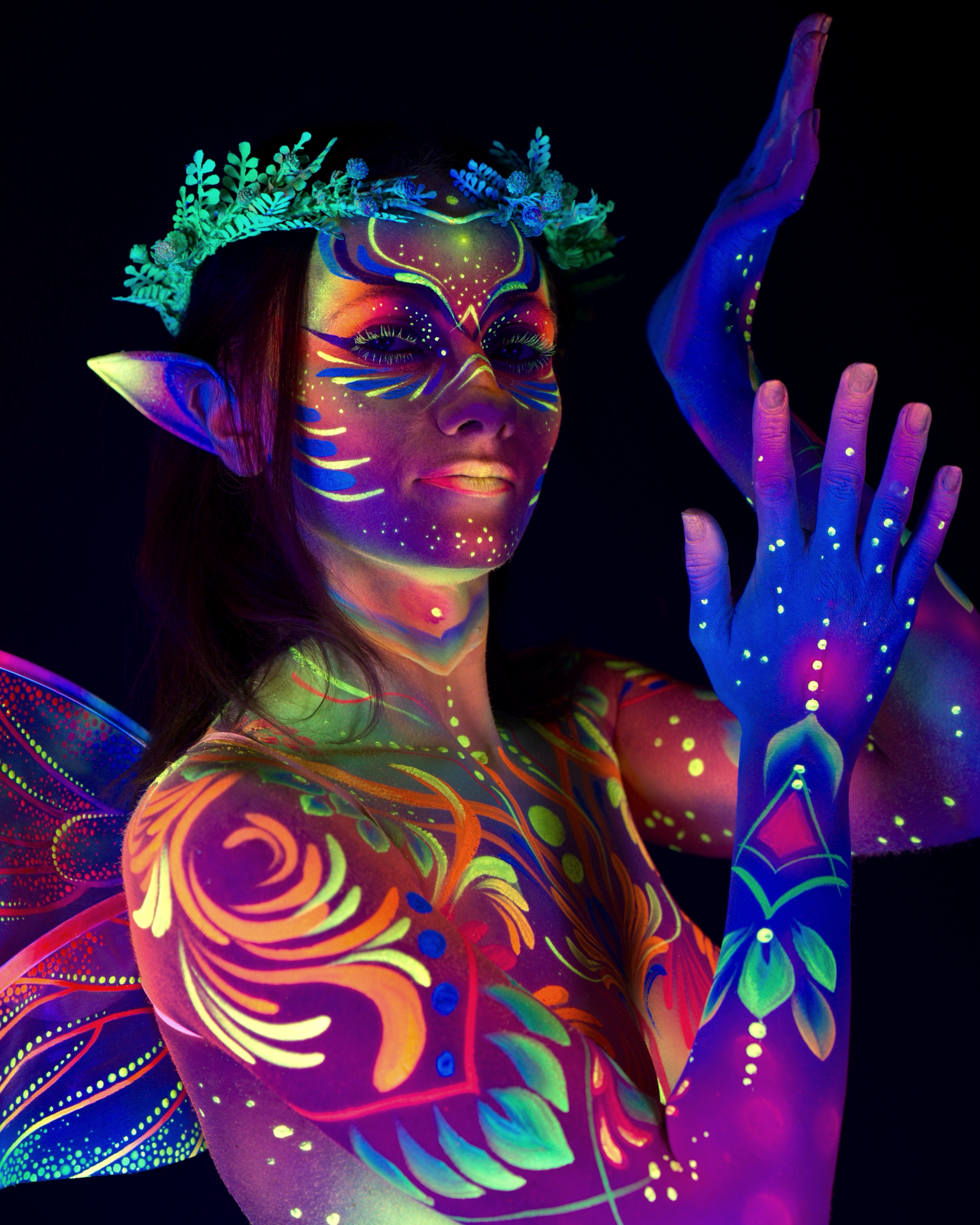 Marina stands in a close three-quarter pose, upper body toward the camera. A fairy themed bodypaint design in orange, pink and green covers her, with a rose pattern on the chest and wing shapes across the shoulders. Bodypaint by Joana Rohr. Photographed by Elad Itzkin Photography under UV blacklight in his studio in Gesundbrunnen, Berlin.