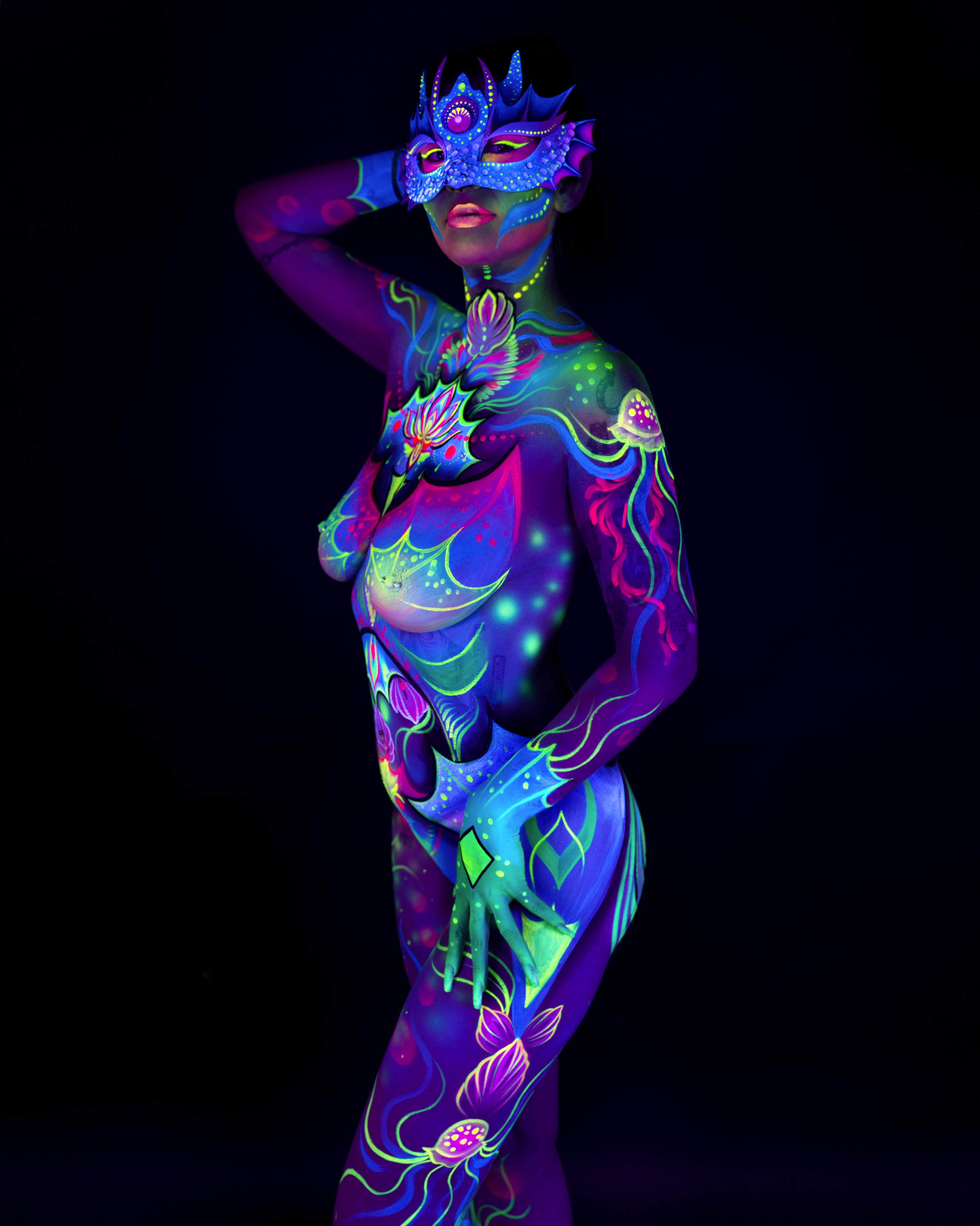 Saskia stands turned slightly toward the camera in a three-quarter pose. The bodypaint is an abstract floral design in blue, purple and pink, with a crowned mask covering the upper face. Bodypaint by Joana Rohr. Photographed by Elad Itzkin Photography under UV blacklight in his studio in Gesundbrunnen, Berlin.