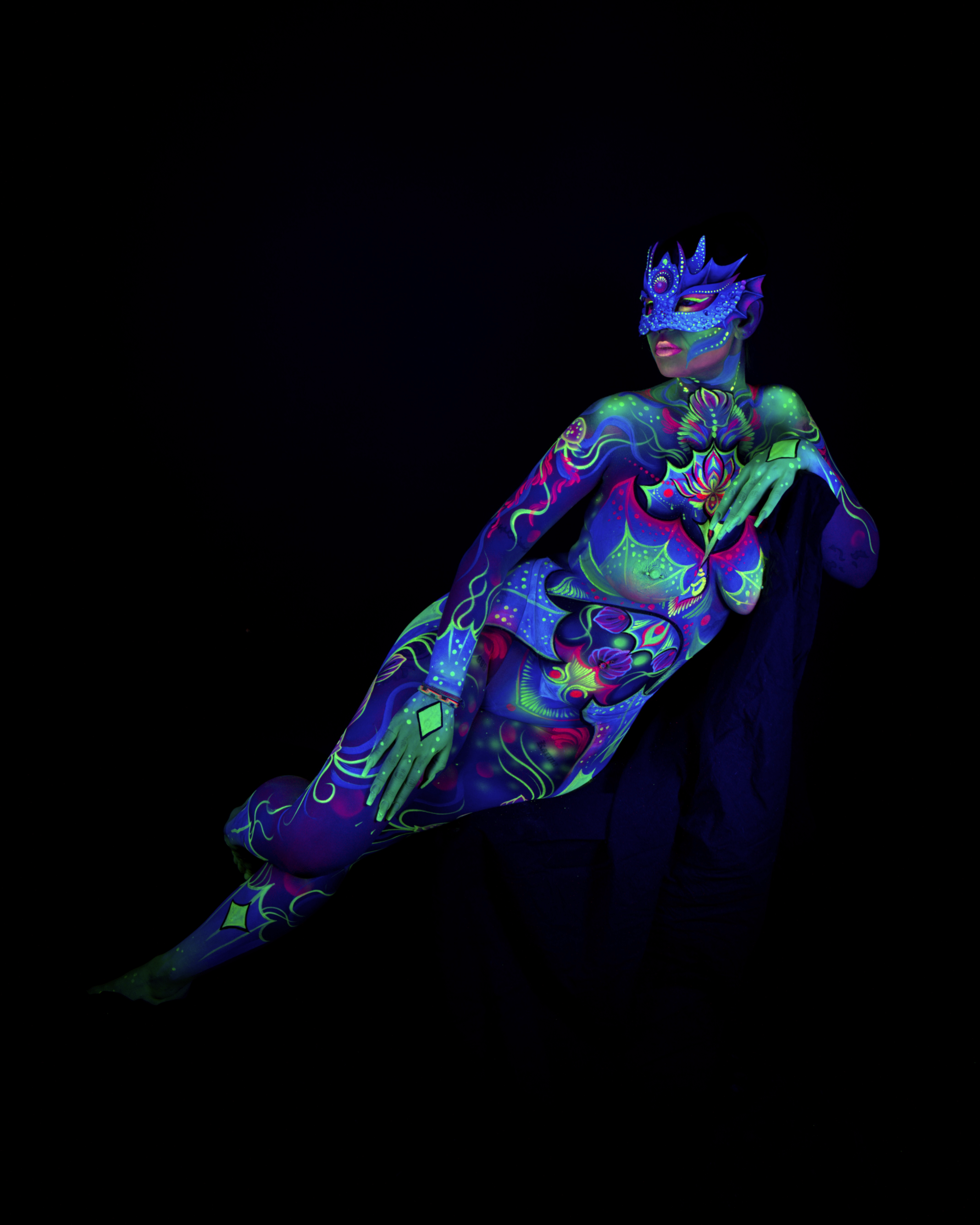 Saskia reclines, leaning back in a three-quarter pose. The bodypaint is an abstract mandala design in blue, purple and pink, with a crowned mask over the eyes and a floral pattern across the torso. Bodypaint by Joana Rohr. Photographed by Elad Itzkin Photography under UV blacklight in his studio in Gesundbrunnen, Berlin.