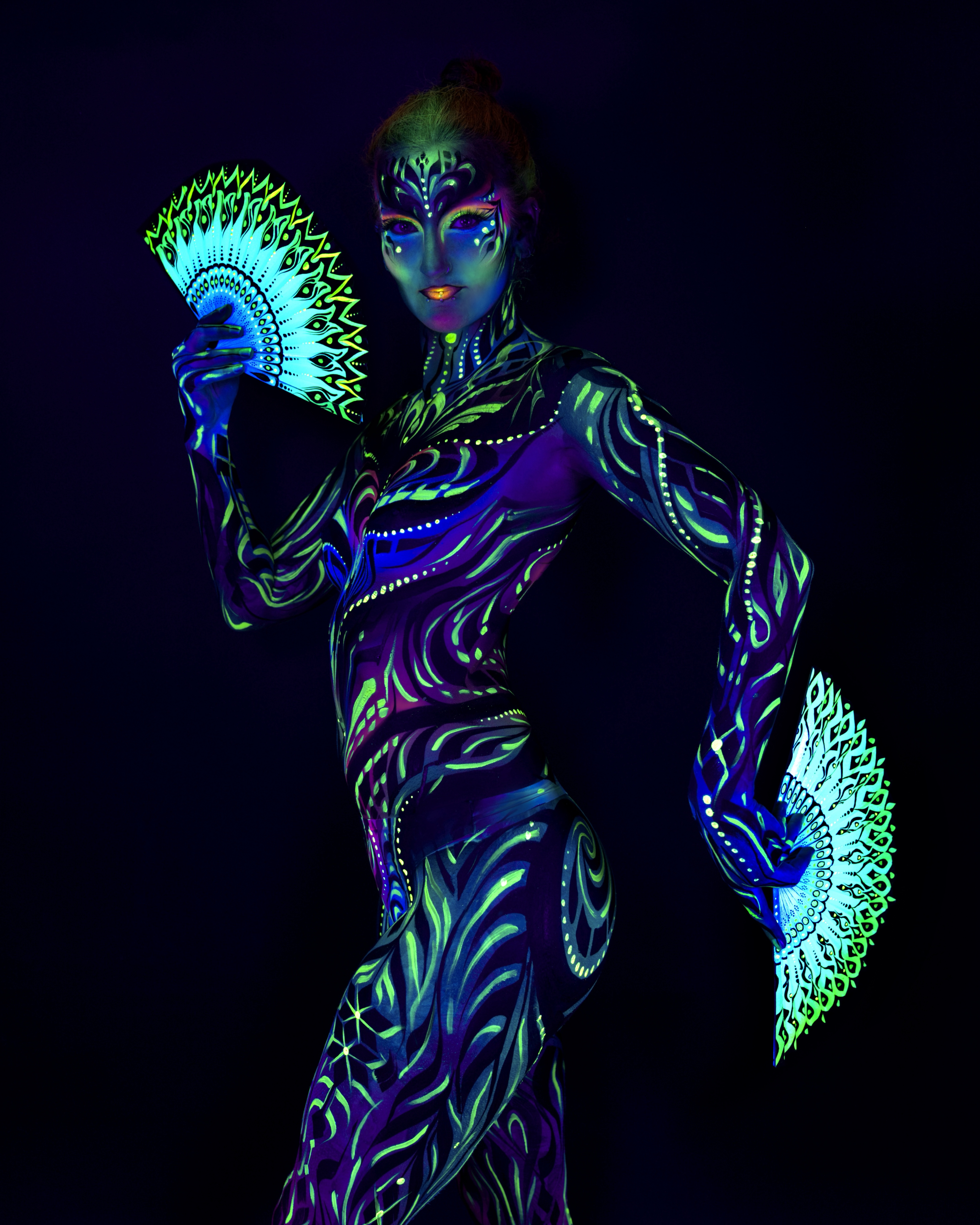 Joey stands in profile, turned toward the camera, with decorative feather-pattern fans in the frame. The bodypaint is a tribal linework design in blue and green across the torso and face. Bodypaint by Joana Rohr and Zami. Photographed by Elad Itzkin Photography under UV blacklight in his studio in Gesundbrunnen, Berlin.