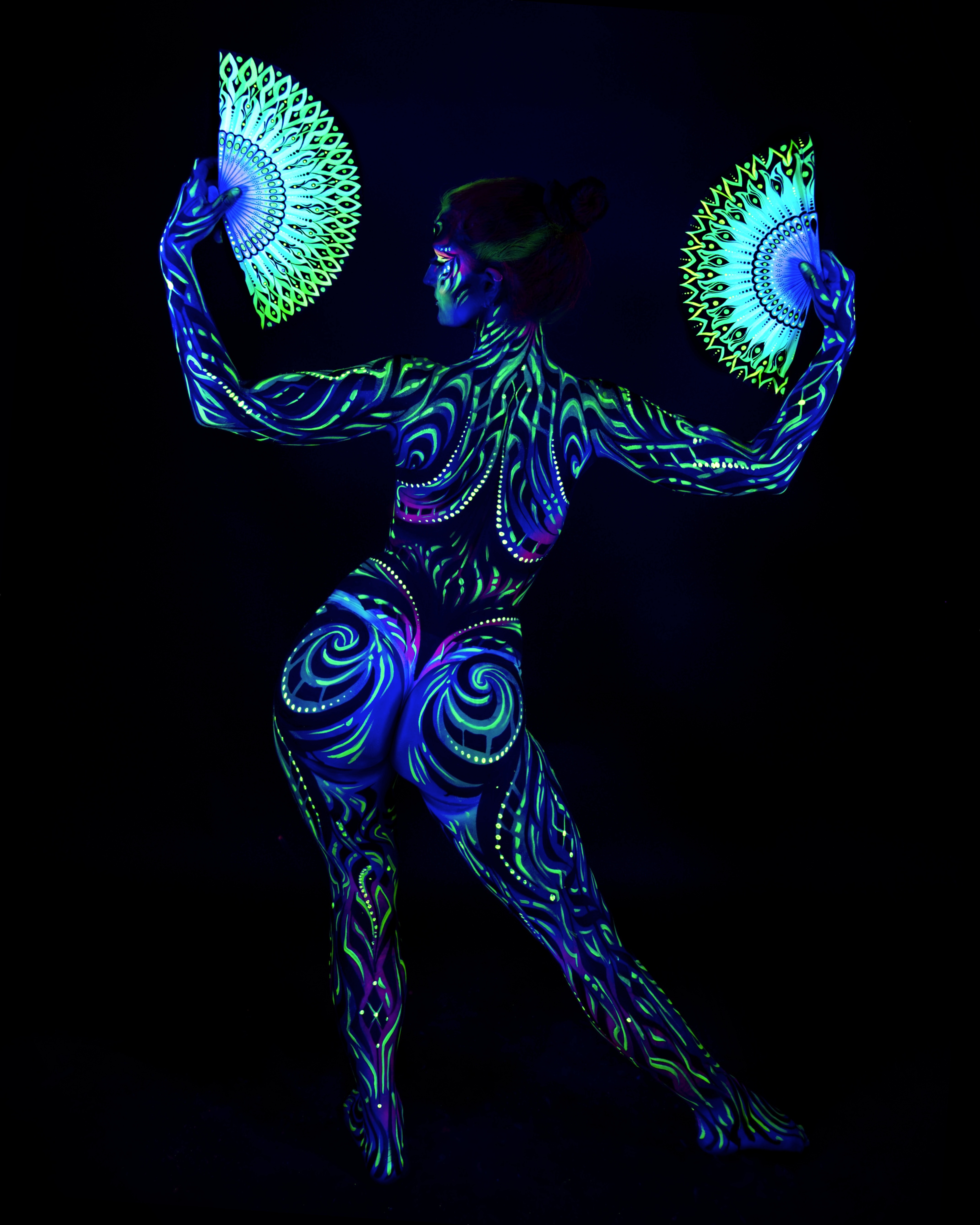 Joey stands with the back to the camera, feather-pattern fans visible above the shoulders. The bodypaint is a tribal linework and swirl design in blue and green, covering the back and lower body. Bodypaint by Joana Rohr and Zami. Photographed by Elad Itzkin Photography under UV blacklight in his studio in Gesundbrunnen, Berlin.