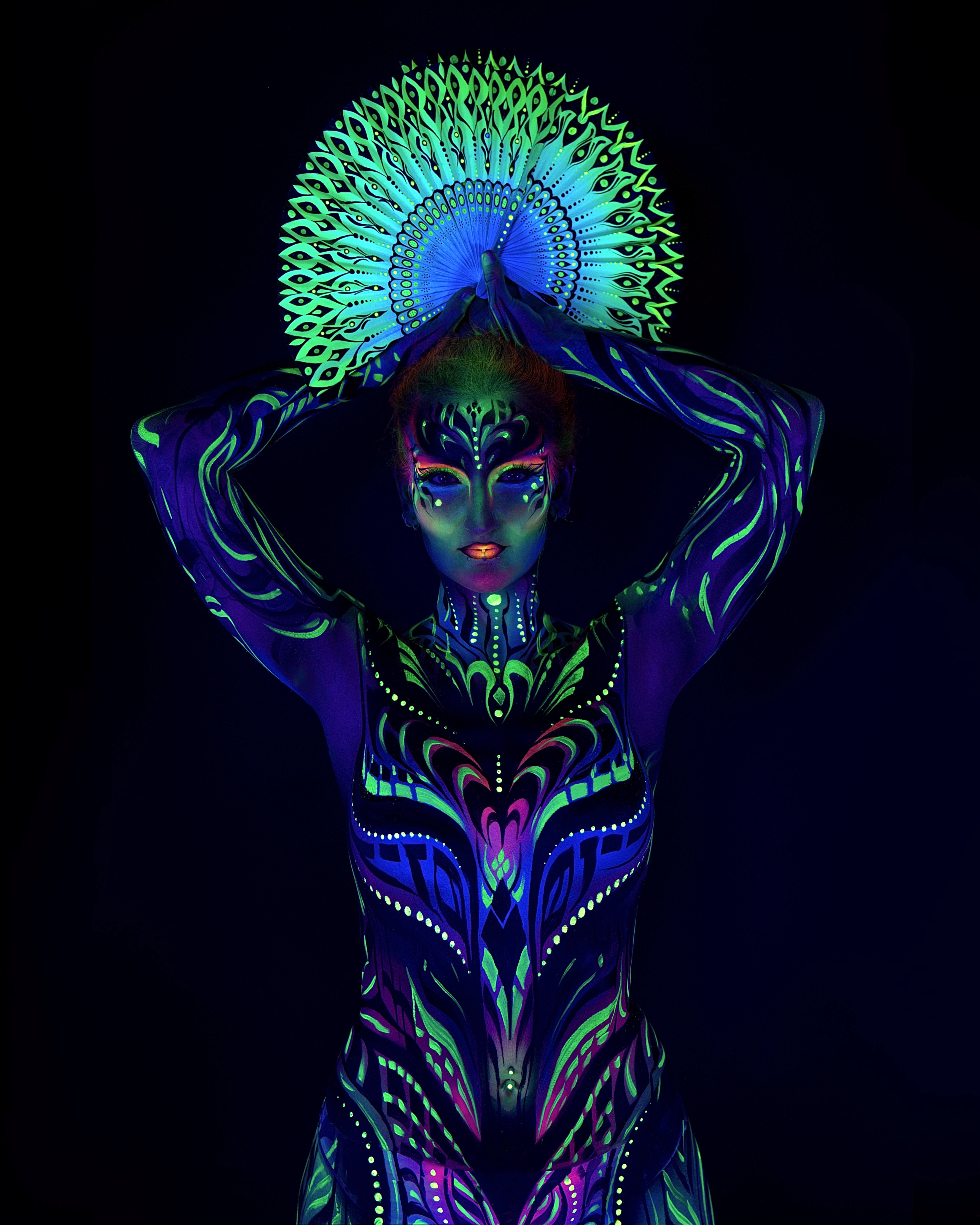 Joey stands facing the camera, a decorative feather-pattern fan raised above the head. The bodypaint is a tribal linework design in blue, green and pink covering the torso. Bodypaint by Joana Rohr and Zami. Photographed by Elad Itzkin Photography under UV blacklight in his studio in Gesundbrunnen, Berlin.