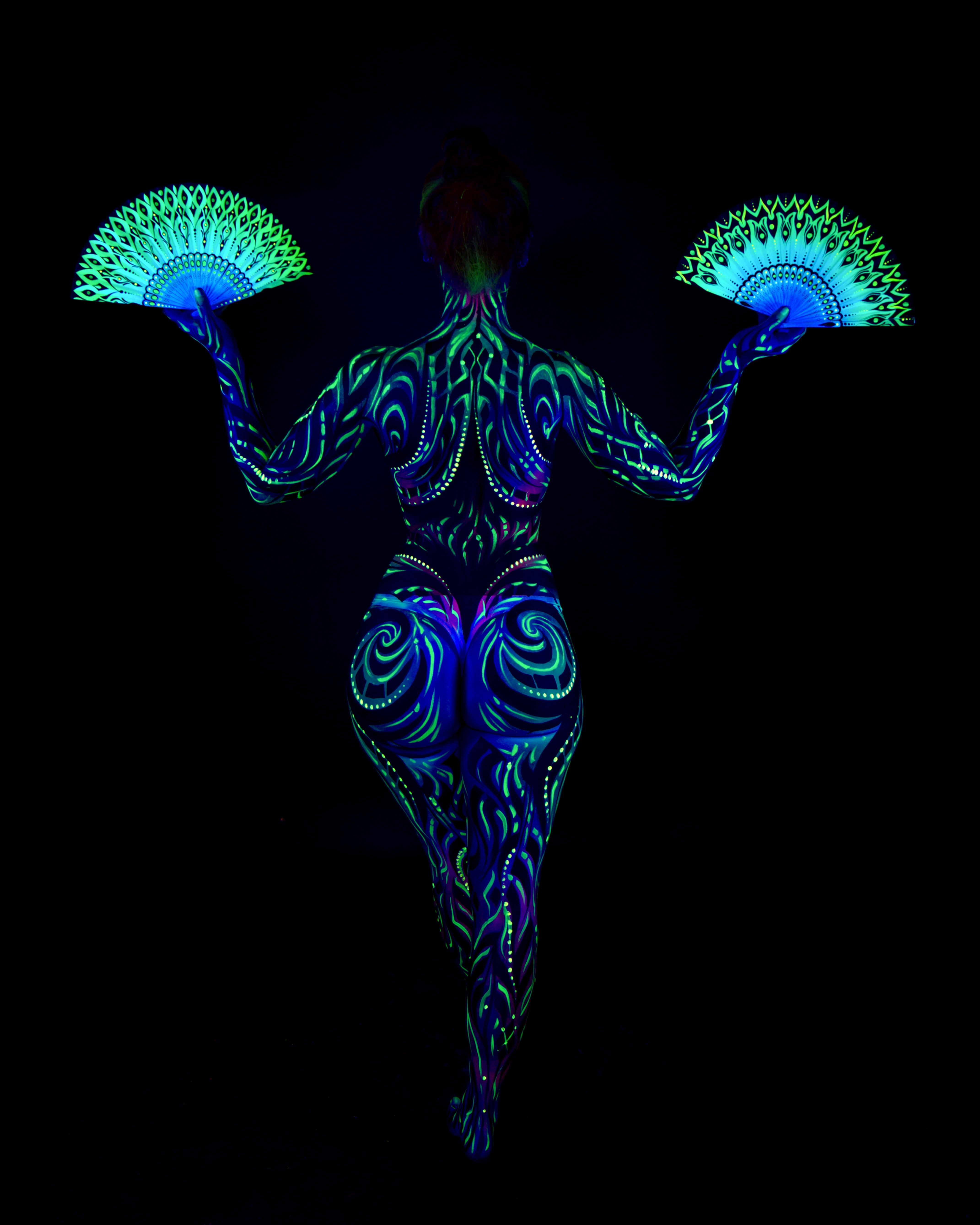 Joey stands with the back to the camera, feather-pattern fans positioned to the sides. The bodypaint is a tribal swirl design in blue and green across the back and lower body. Bodypaint by Joana Rohr and Zami. Photographed by Elad Itzkin Photography under UV blacklight in his studio in Gesundbrunnen, Berlin.