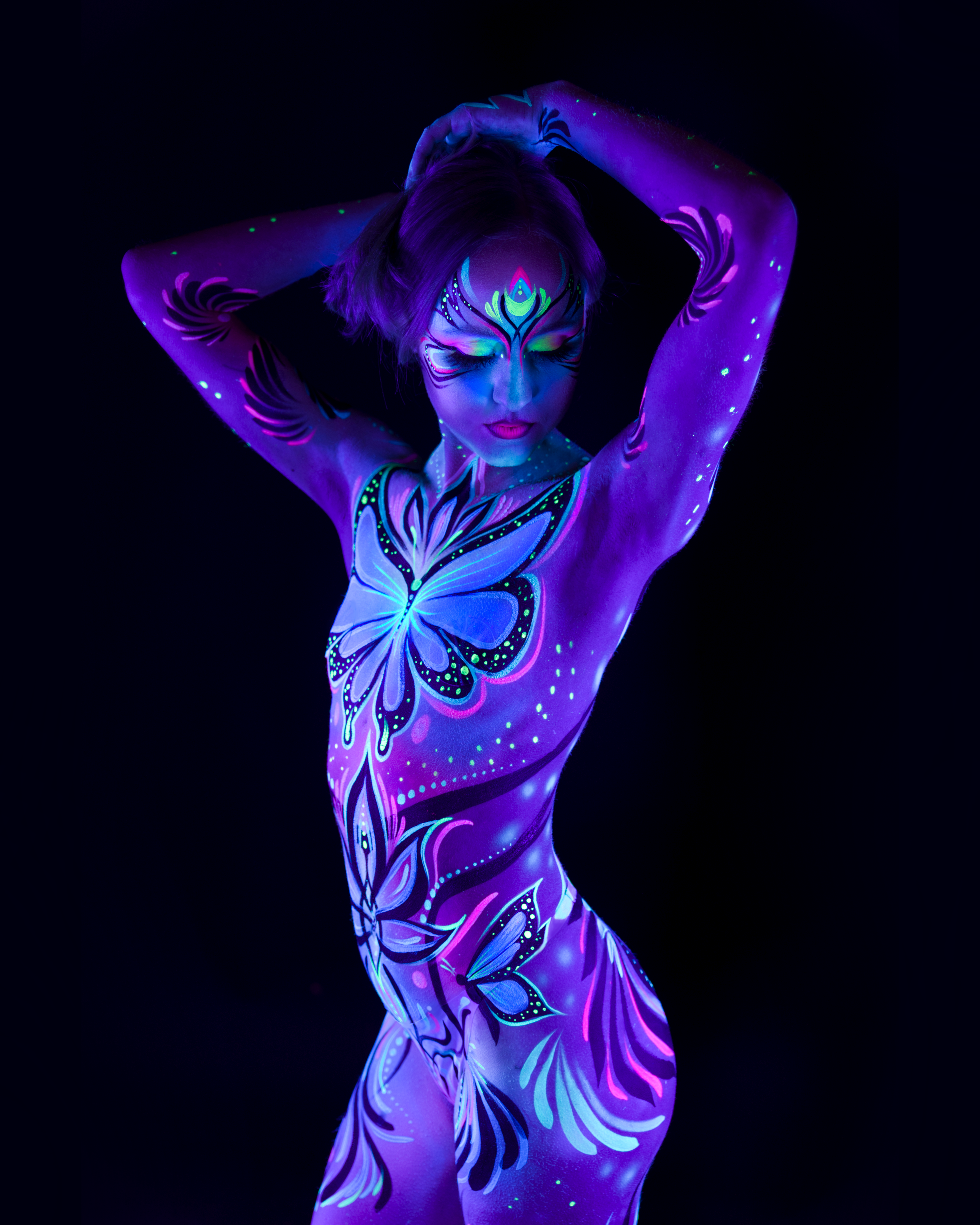 Livia stands facing the camera in a frontal pose, head tilted slightly down. The bodypaint is a butterfly and floral linework design in purple, blue and pink covering the chest and torso. Bodypaint by Joana Rohr. Photographed by Elad Itzkin Photography under UV blacklight in his studio in Gesundbrunnen, Berlin.
