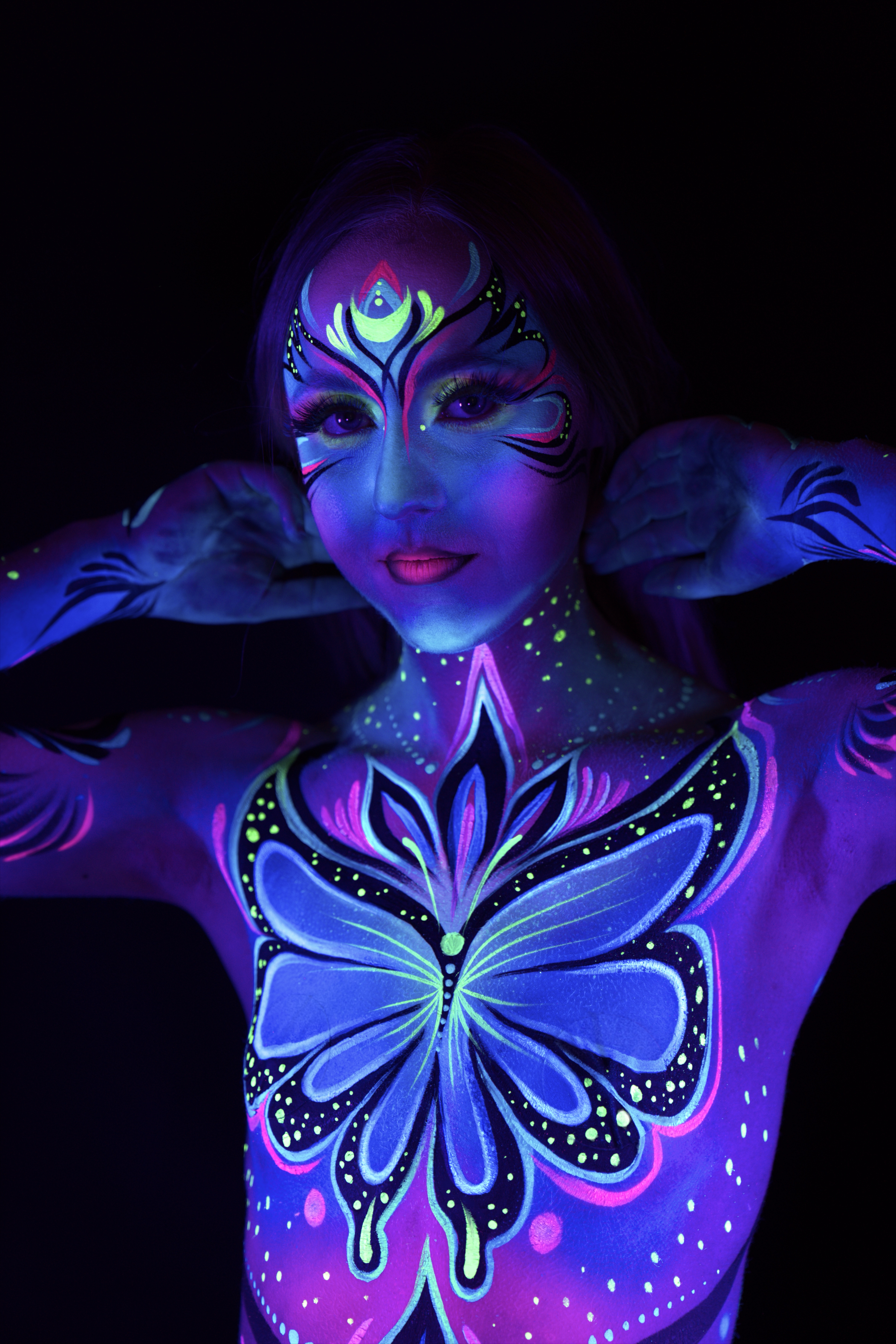 Livia stands in a close three-quarter pose, facing the camera. The bodypaint features a large butterfly motif in blue and purple on the chest, part of a wider floral design across the torso. Bodypaint by Joana Rohr. Photographed by Elad Itzkin Photography under UV blacklight in his studio in Gesundbrunnen, Berlin.