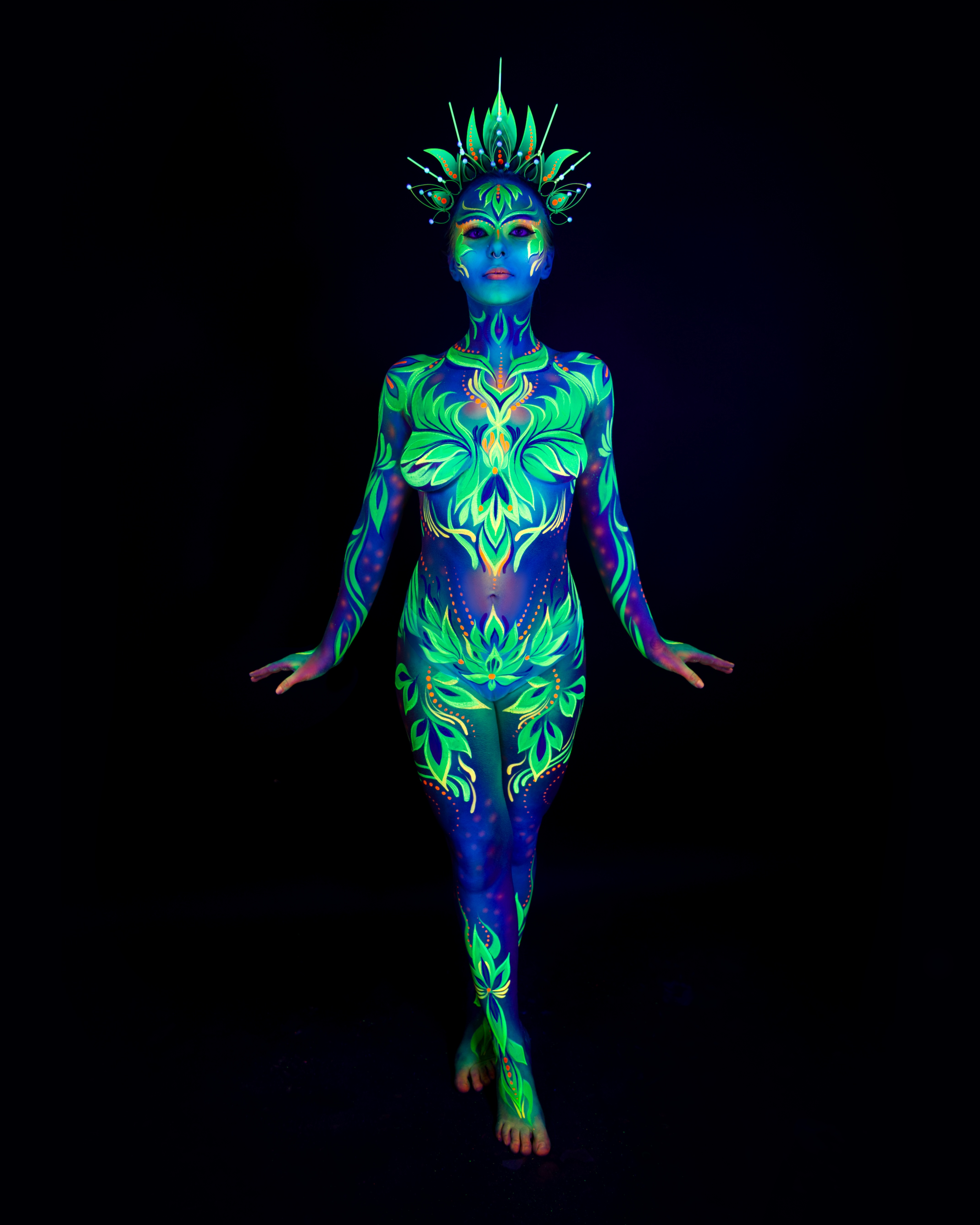Katrina K stands facing the camera in a walking pose, wearing a leaf-shaped crown headpiece. The bodypaint is a green and blue leaf and floral design with orange accents, covering the body. Bodypaint by Joana Rohr. Photographed by Elad Itzkin Photography under UV blacklight in his studio in Gesundbrunnen, Berlin.
