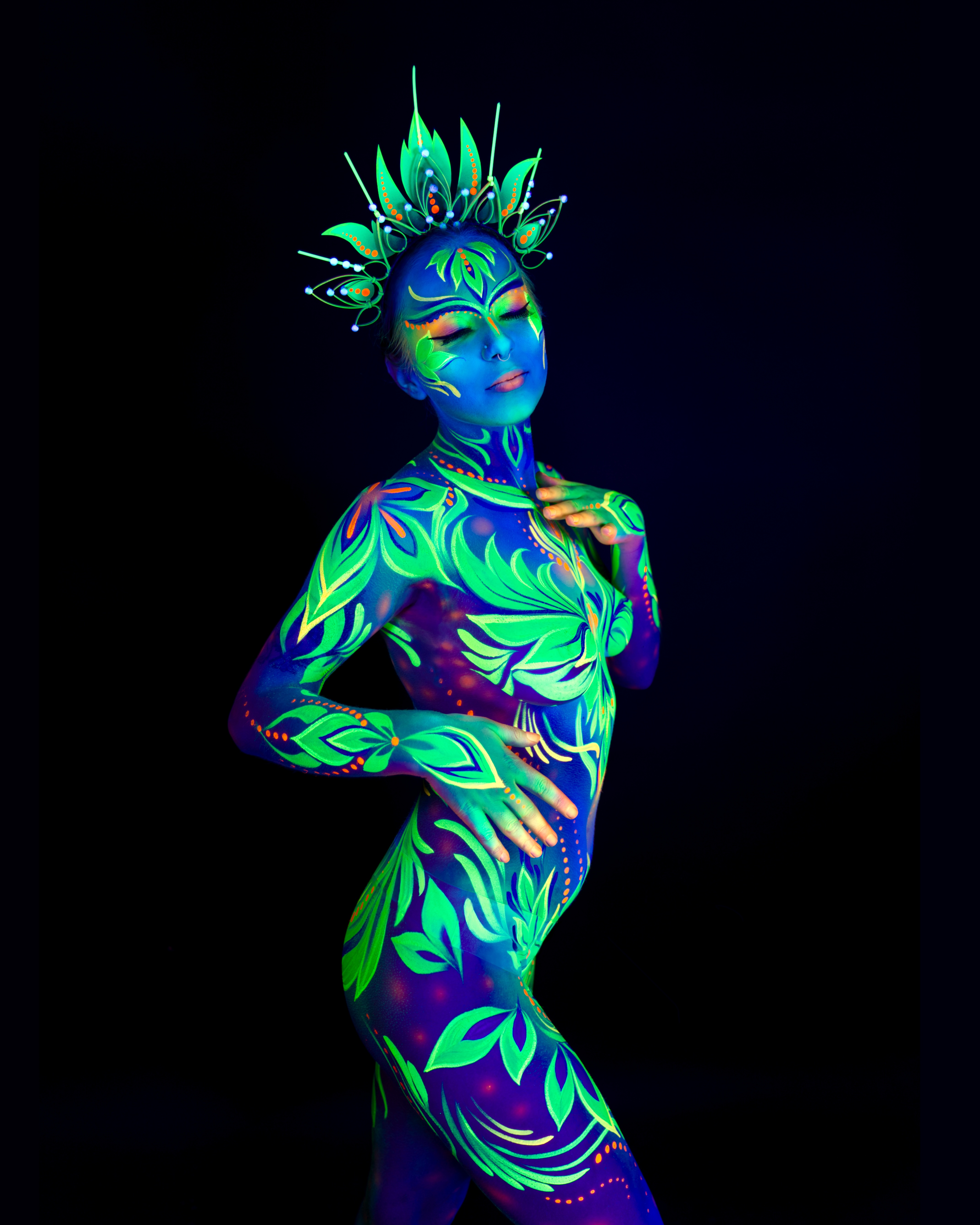 Katrina K stands in a three-quarter pose wearing an ornate crown-like headpiece. Her body is painted in a blue base with bright green leaf and floral patterns. Bodypaint by Joana Rohr. Photographed by Elad Itzkin Photography under UV blacklight in his studio in Gesundbrunnen, Berlin.