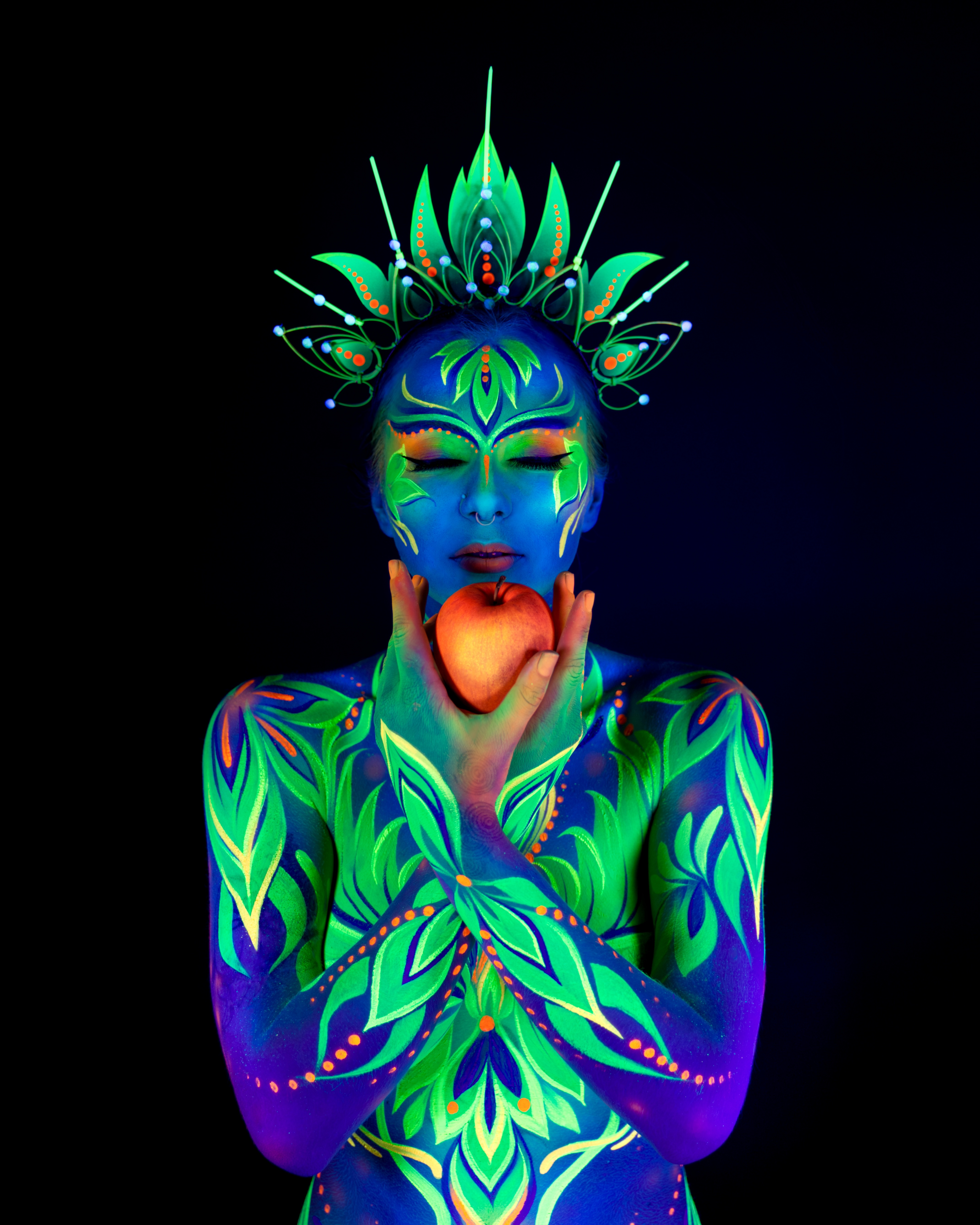 Katrina K stands facing the camera in an ornate crown-like headpiece. Her body is painted with a green and blue floral leaf pattern accented with orange dots. Bodypaint by Joana Rohr. Photographed by Elad Itzkin Photography under UV blacklight in his studio in Gesundbrunnen, Berlin.