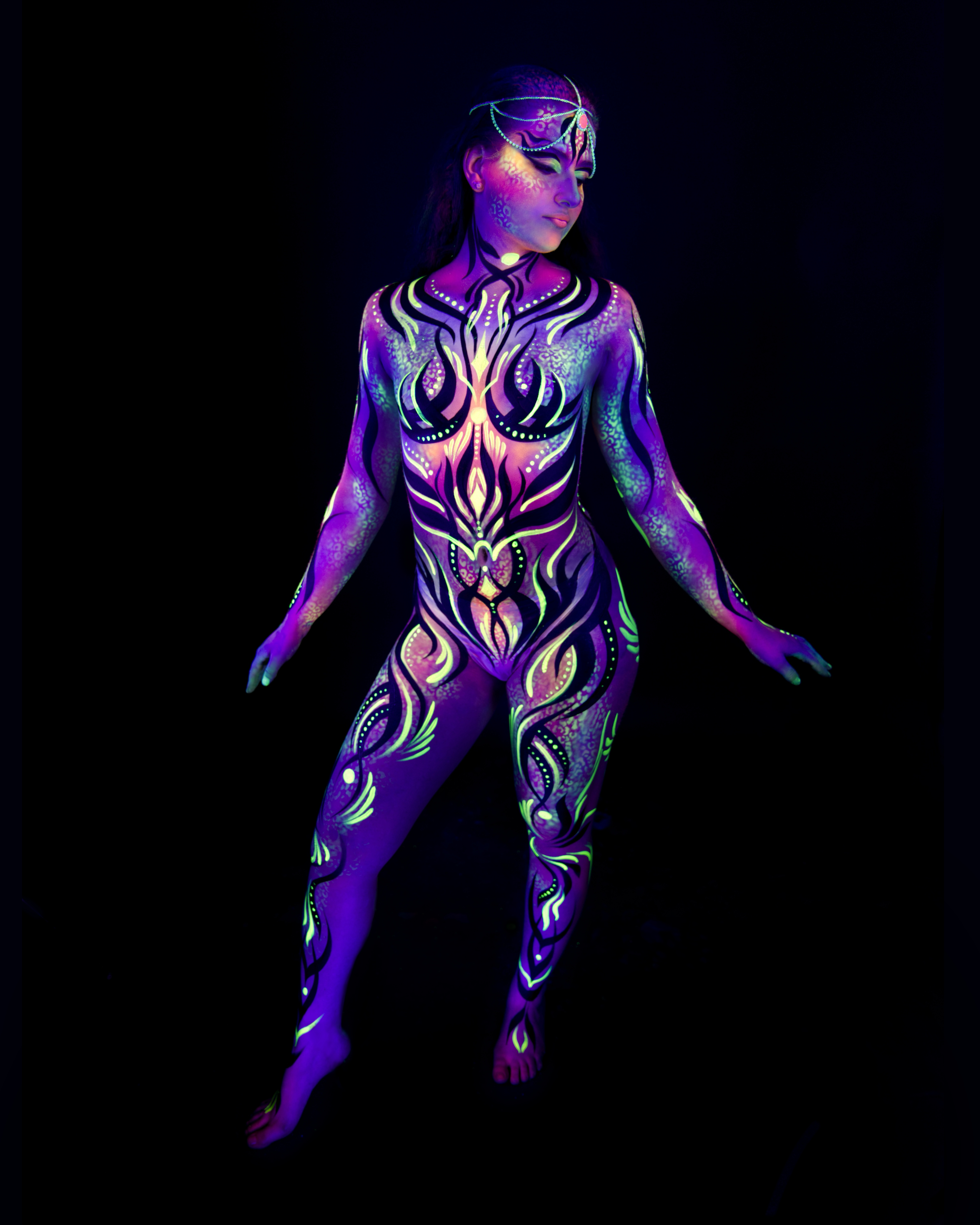 Elizaveta stands in a slight side-facing pose. Her body is painted in a purple and pink abstract linework design with flame-like and floral motifs. Bodypaint by Joana Rohr. Photographed by Elad Itzkin Photography under UV blacklight in his studio in Gesundbrunnen, Berlin.