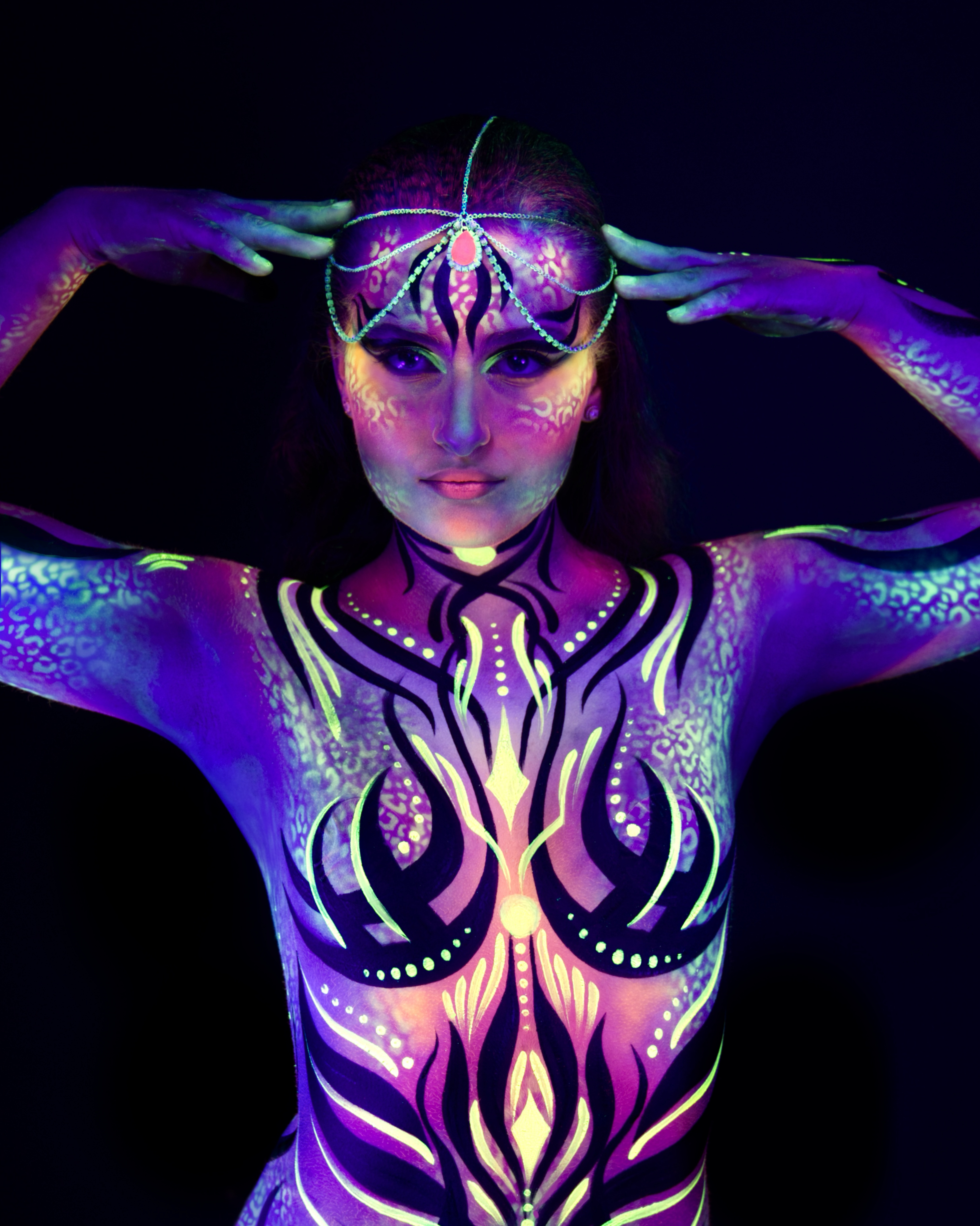 Elizaveta stands facing the camera, shown from the head to the chest, wearing a chain-style headpiece. Her body is painted in a purple, orange, and yellow abstract flame pattern. Bodypaint by Joana Rohr. Photographed by Elad Itzkin Photography under UV blacklight in his studio in Gesundbrunnen, Berlin.