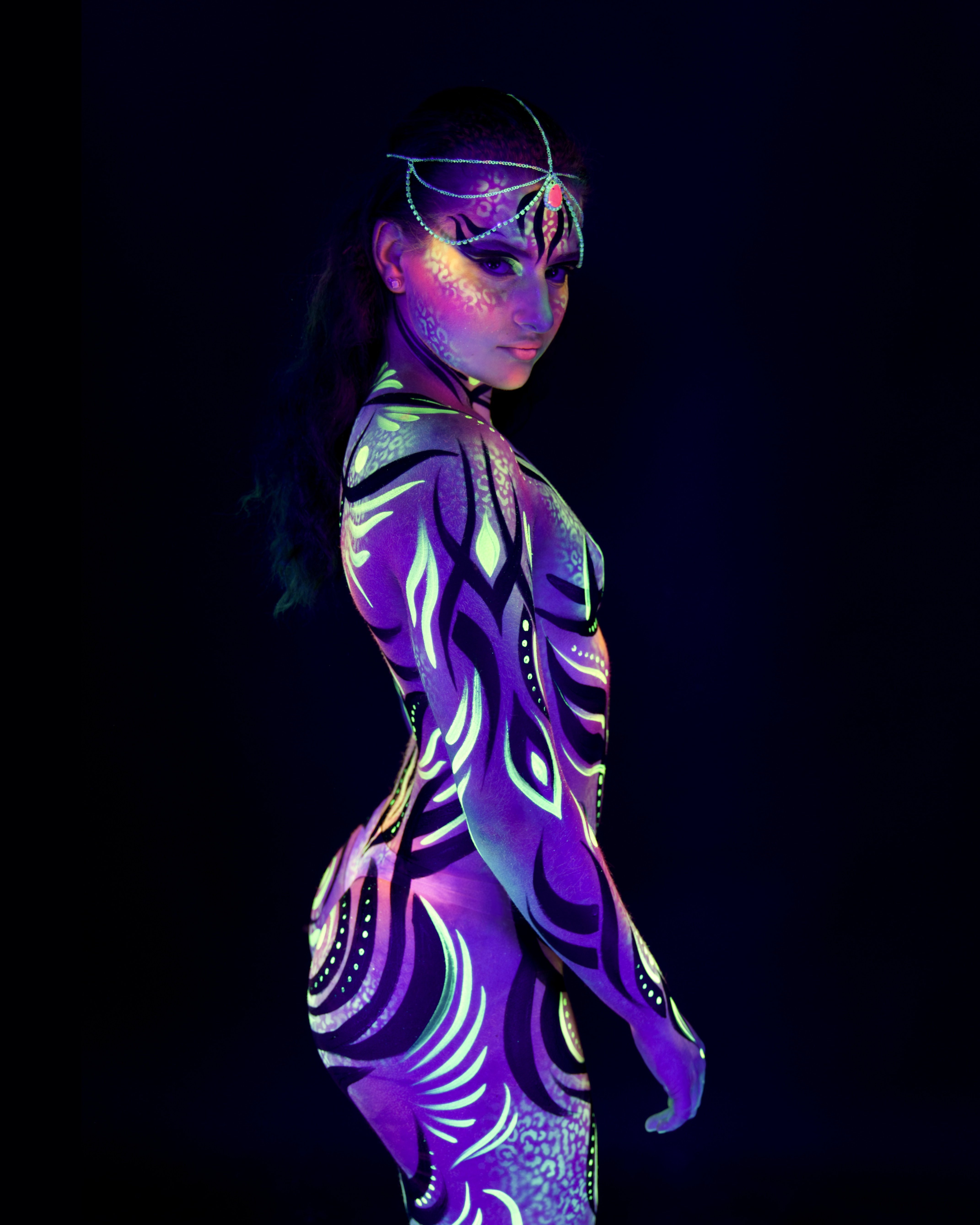 Elizaveta stands in profile, turned to one side. Her body is painted in a purple abstract design with geometric diamond and wave-like linework. Bodypaint by Joana Rohr. Photographed by Elad Itzkin Photography under UV blacklight in his studio in Gesundbrunnen, Berlin.