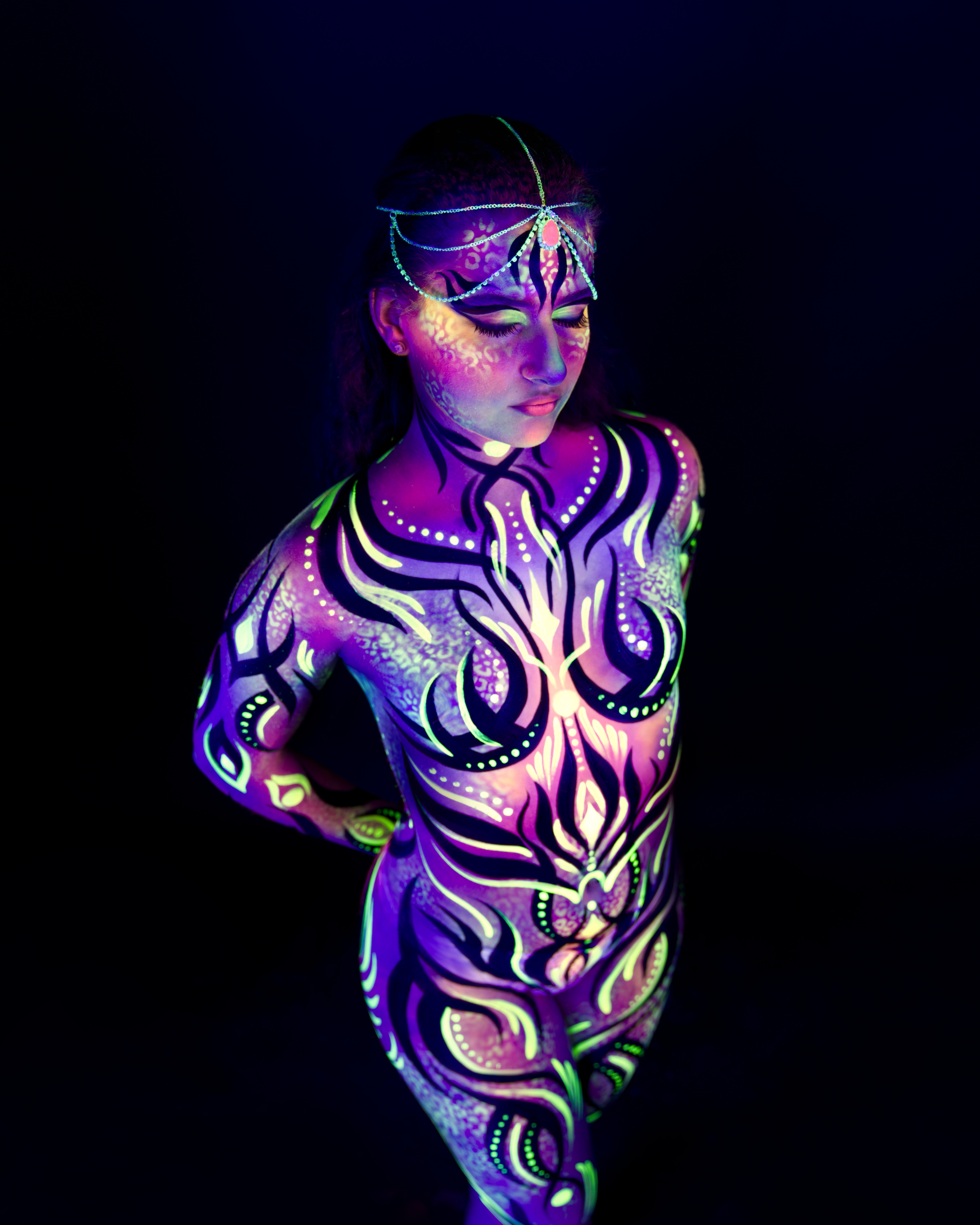 Elizaveta stands in a three-quarter pose wearing a chain-style headpiece. Her body is painted in a purple and green abstract flame and linework design. Bodypaint by Joana Rohr. Photographed by Elad Itzkin Photography under UV blacklight in his studio in Gesundbrunnen, Berlin.