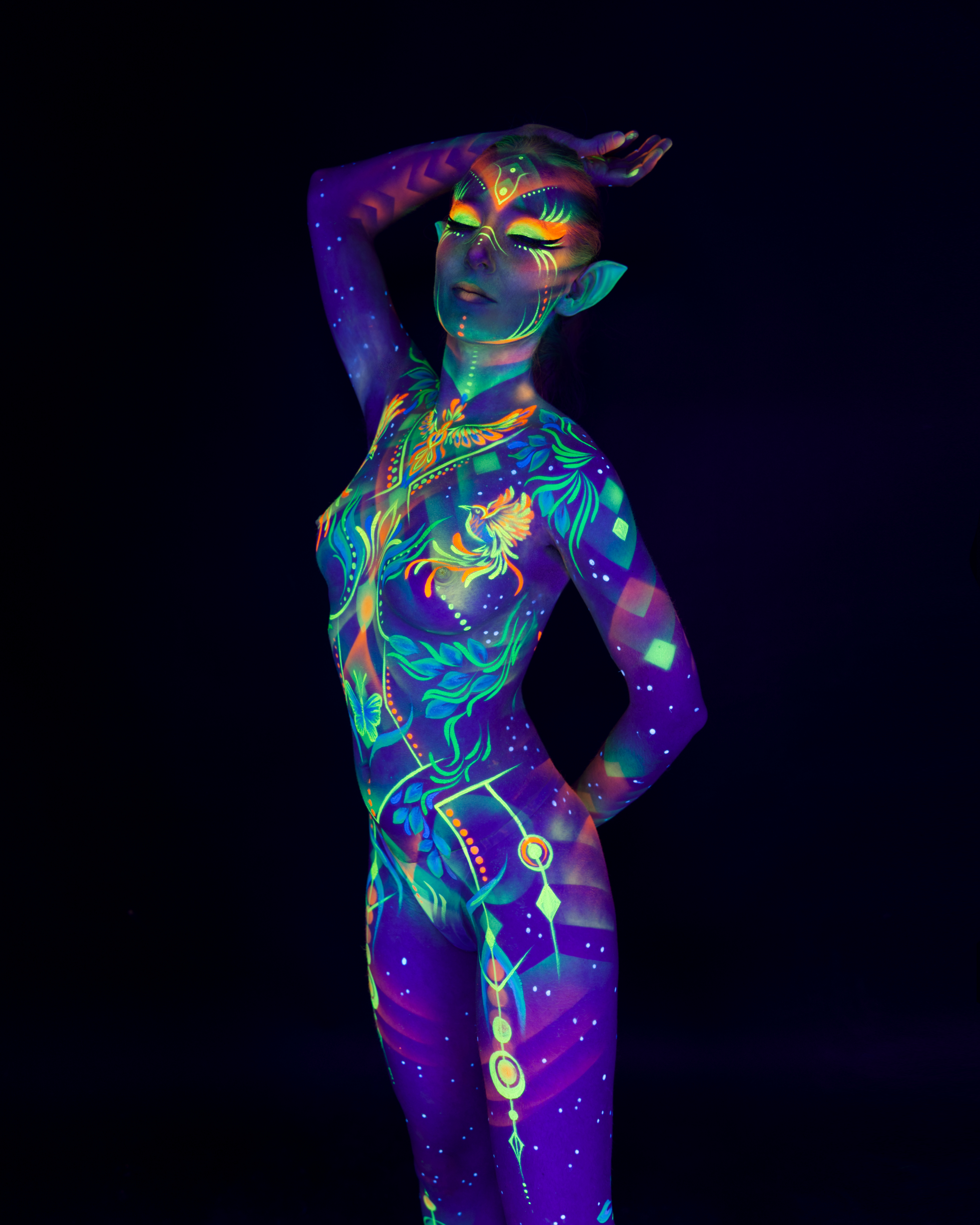 Alena stands in a three-quarter pose with prosthetic pointed ears. Her body is painted in a blue and green design featuring a bird motif and scattered geometric diamond patches in orange and pink. Bodypaint by Joana Rohr. Photographed by Elad Itzkin Photography under UV blacklight in his studio in Gesundbrunnen, Berlin.