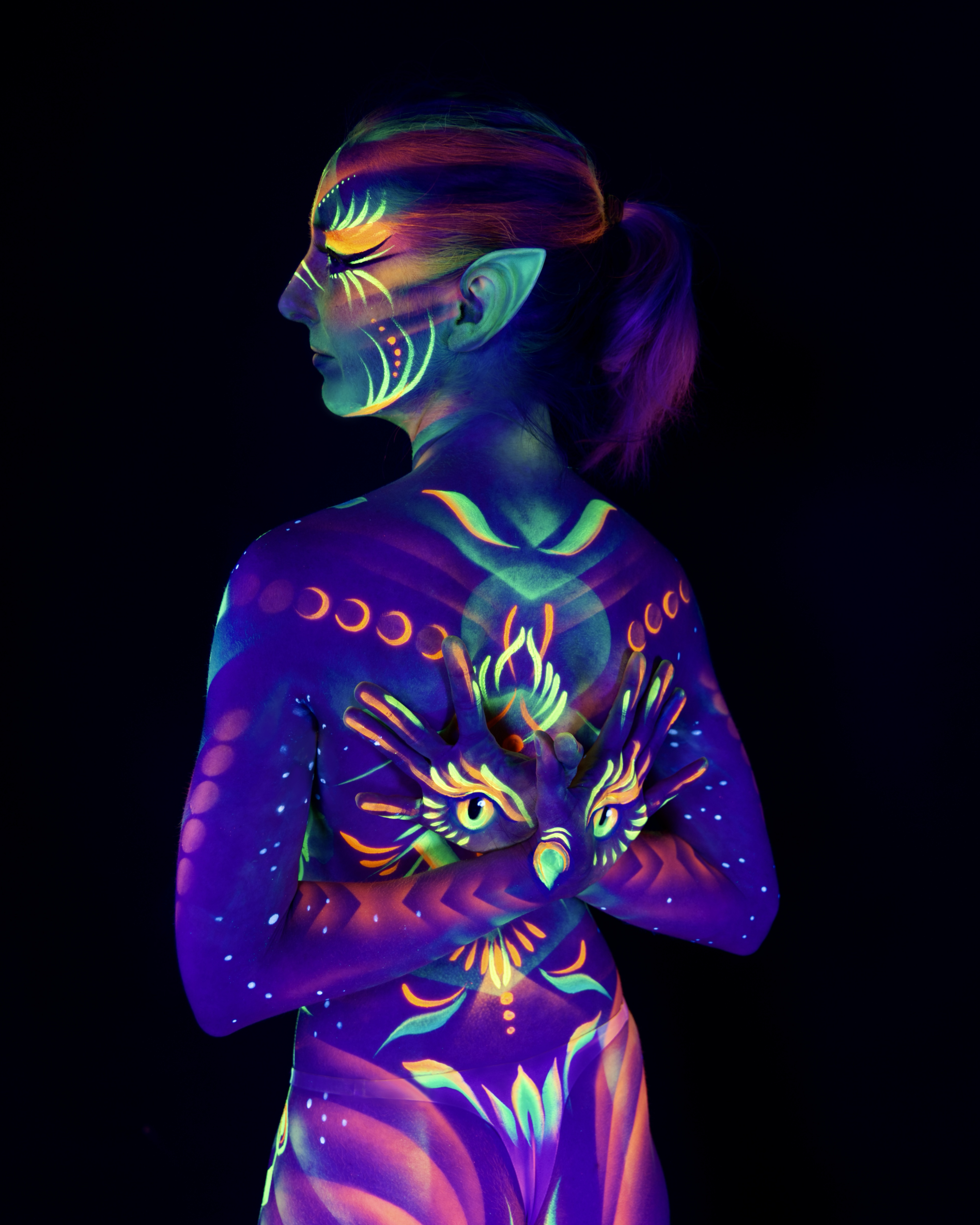 Alena is shown from behind with her face in profile, wearing pointed prosthetic ears. Her back is painted with a bird motif in orange and green, framed by a row of moon phase symbols in purple. Bodypaint by Joana Rohr. Photographed by Elad Itzkin Photography under UV blacklight in his studio in Gesundbrunnen, Berlin.