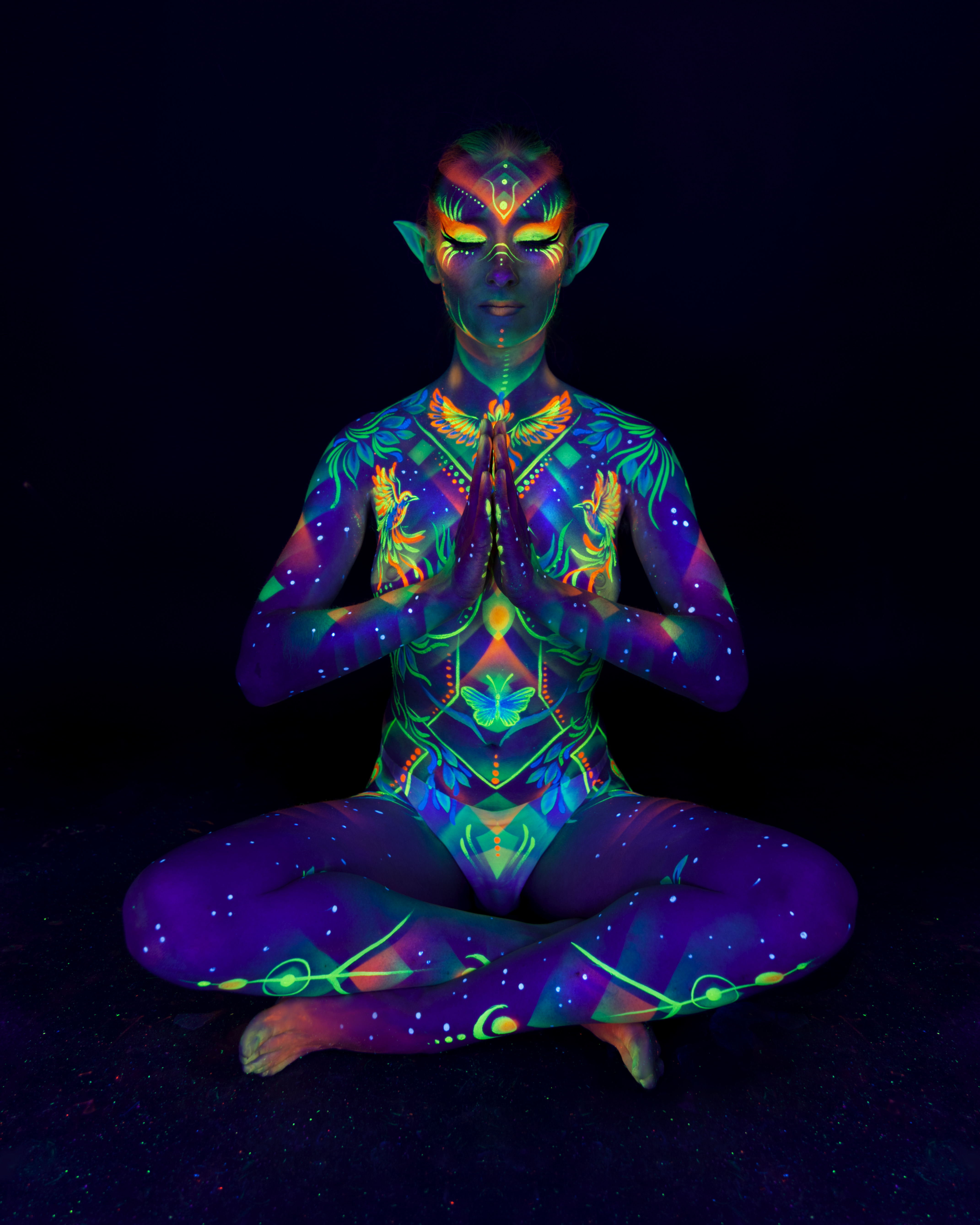 Alena sits cross-legged in a meditative pose, wearing pointed prosthetic ears. Her body is painted with a symmetrical design featuring bird and butterfly motifs in green, orange, and blue. Bodypaint by Joana Rohr. Photographed by Elad Itzkin Photography under UV blacklight in his studio in Gesundbrunnen, Berlin.