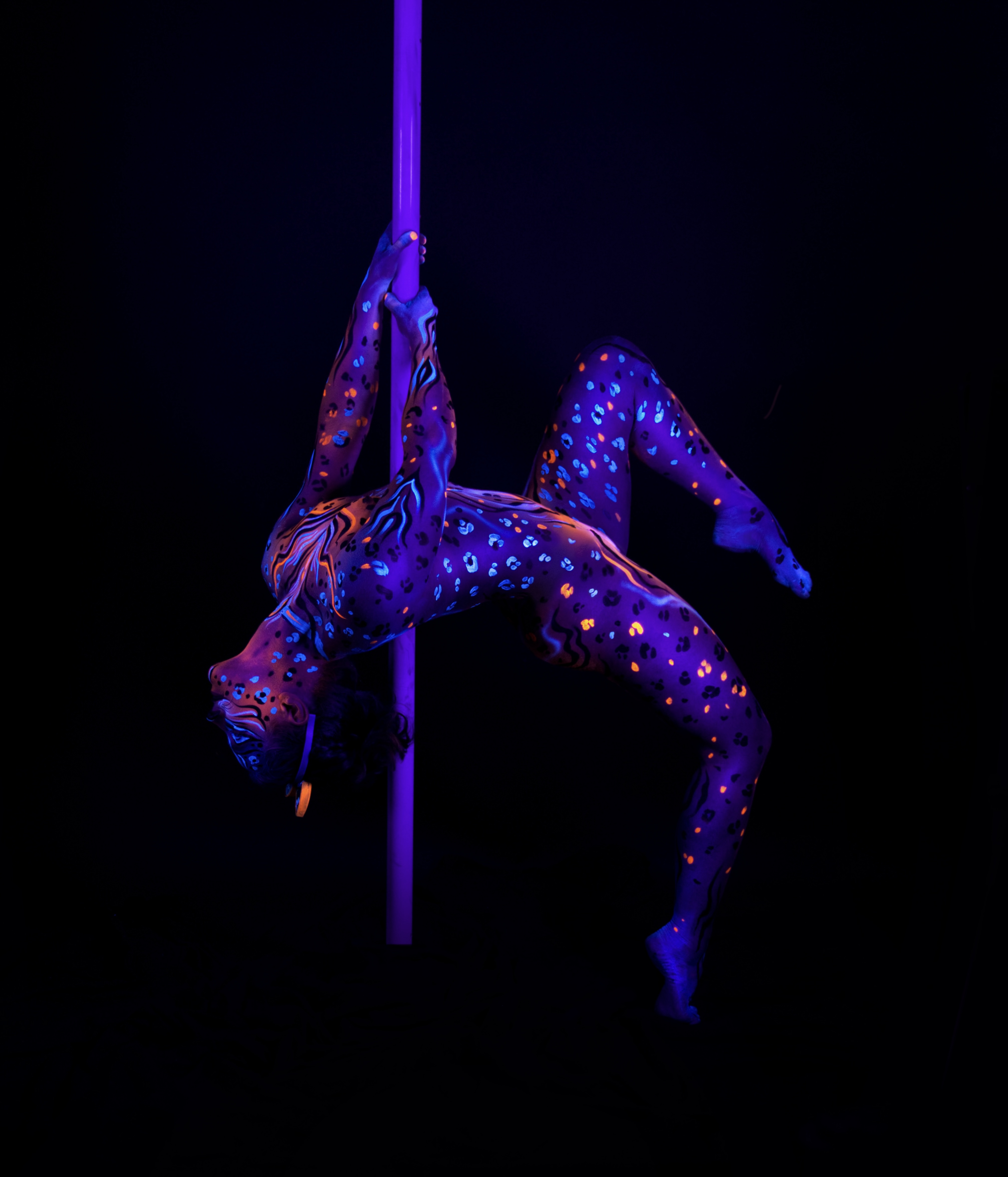 Marcela arches backward while suspended on a pole. Her body is painted with a purple and blue leopard-spot pattern accented with orange highlights. Bodypaint by Joana Rohr, Julia. Photographed by Elad Itzkin Photography under UV blacklight in his studio in Gesundbrunnen, Berlin.