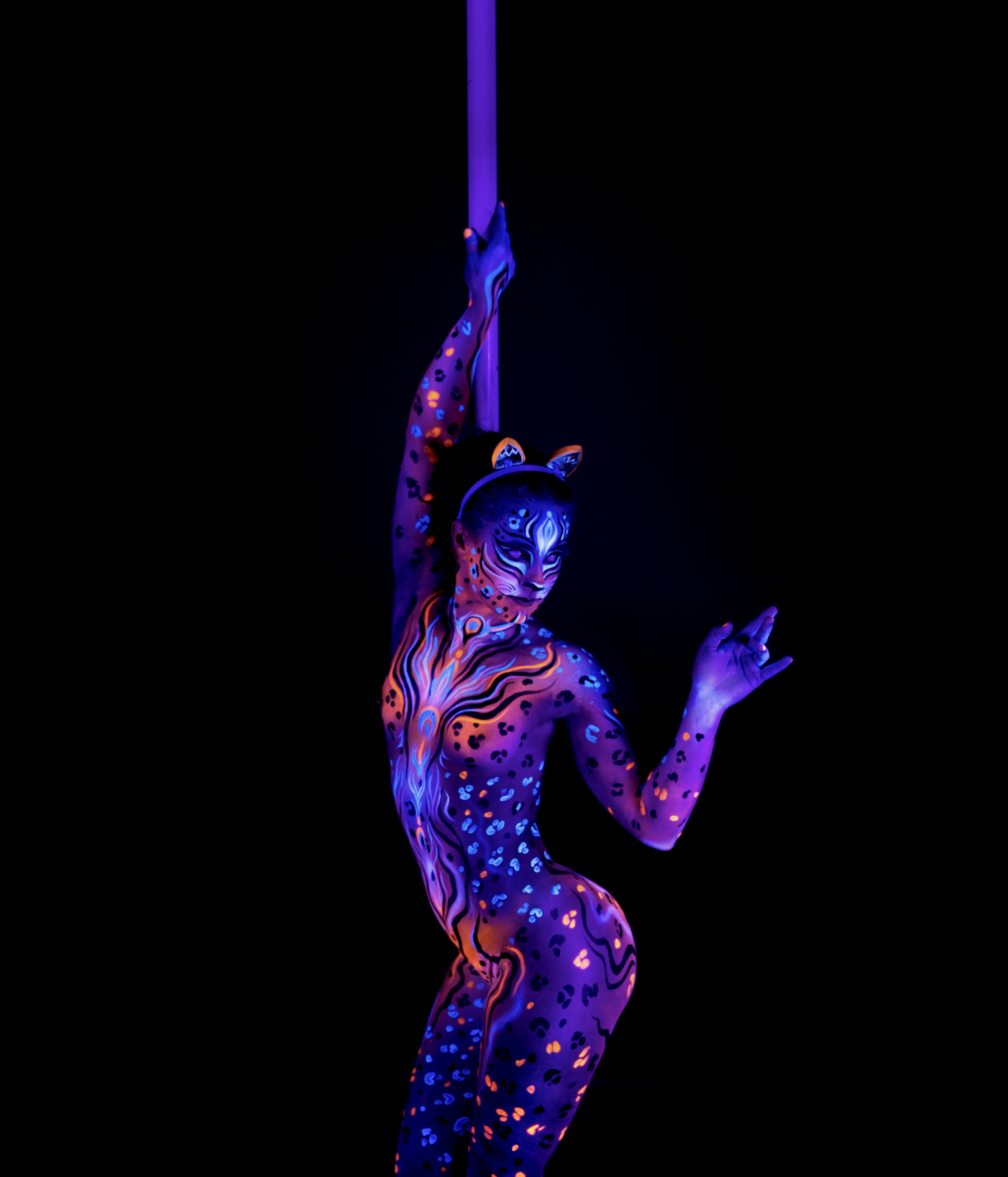 Marcela stands beside a pole with her back turned toward the camera, wearing cat-ear prosthetics. Her body is painted with a purple and orange leopard-spot pattern. Bodypaint by Joana Rohr, Julia. Photographed by Elad Itzkin Photography under UV blacklight in his studio in Gesundbrunnen, Berlin.