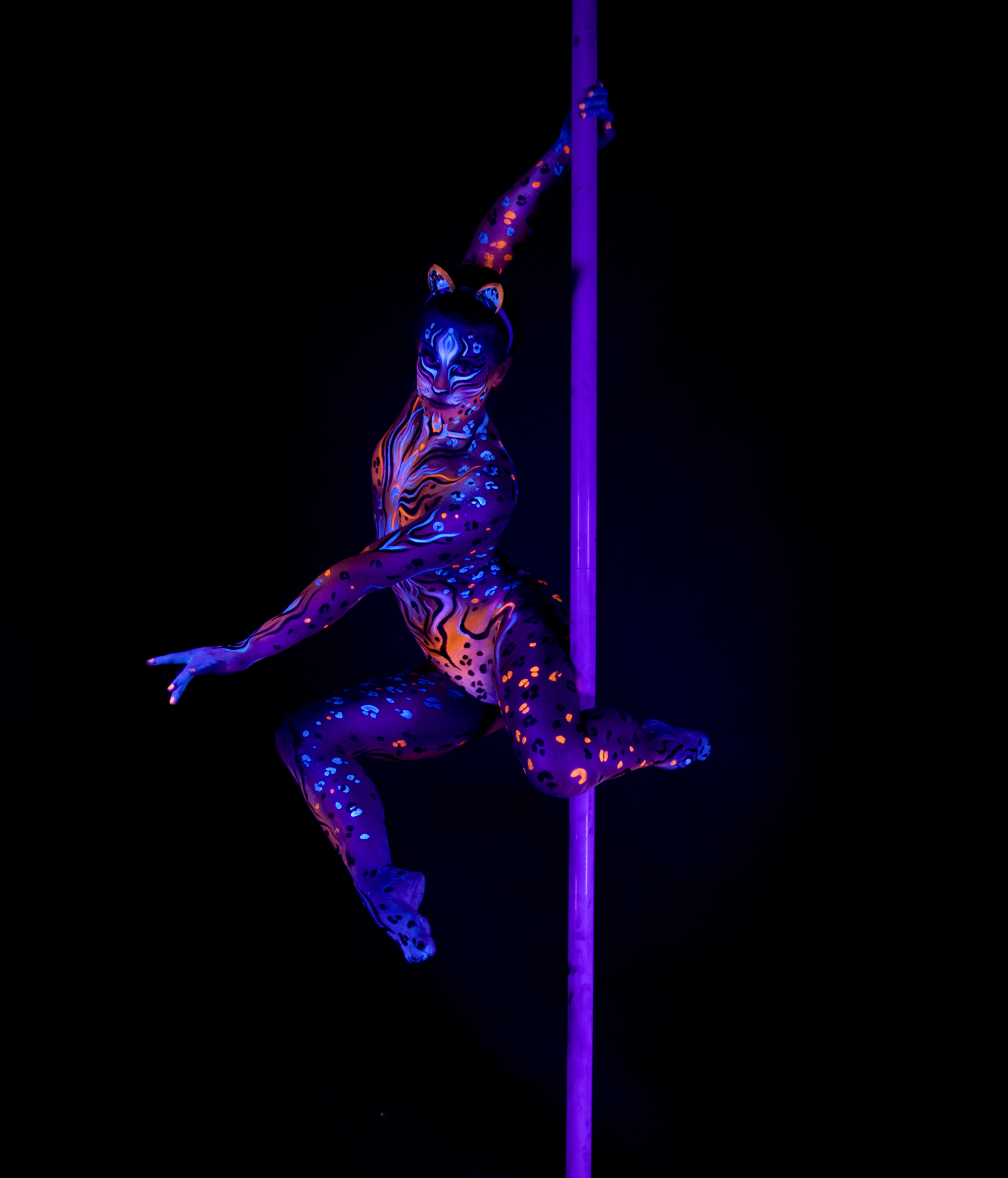 Marcela is suspended in mid-air beside a pole in an acrobatic pose with legs bent, wearing cat-ear prosthetics. Her body is painted with an orange and blue leopard-spot pattern over a purple base. Bodypaint by Joana Rohr, Julia. Photographed by Elad Itzkin Photography under UV blacklight in his studio in Gesundbrunnen, Berlin.