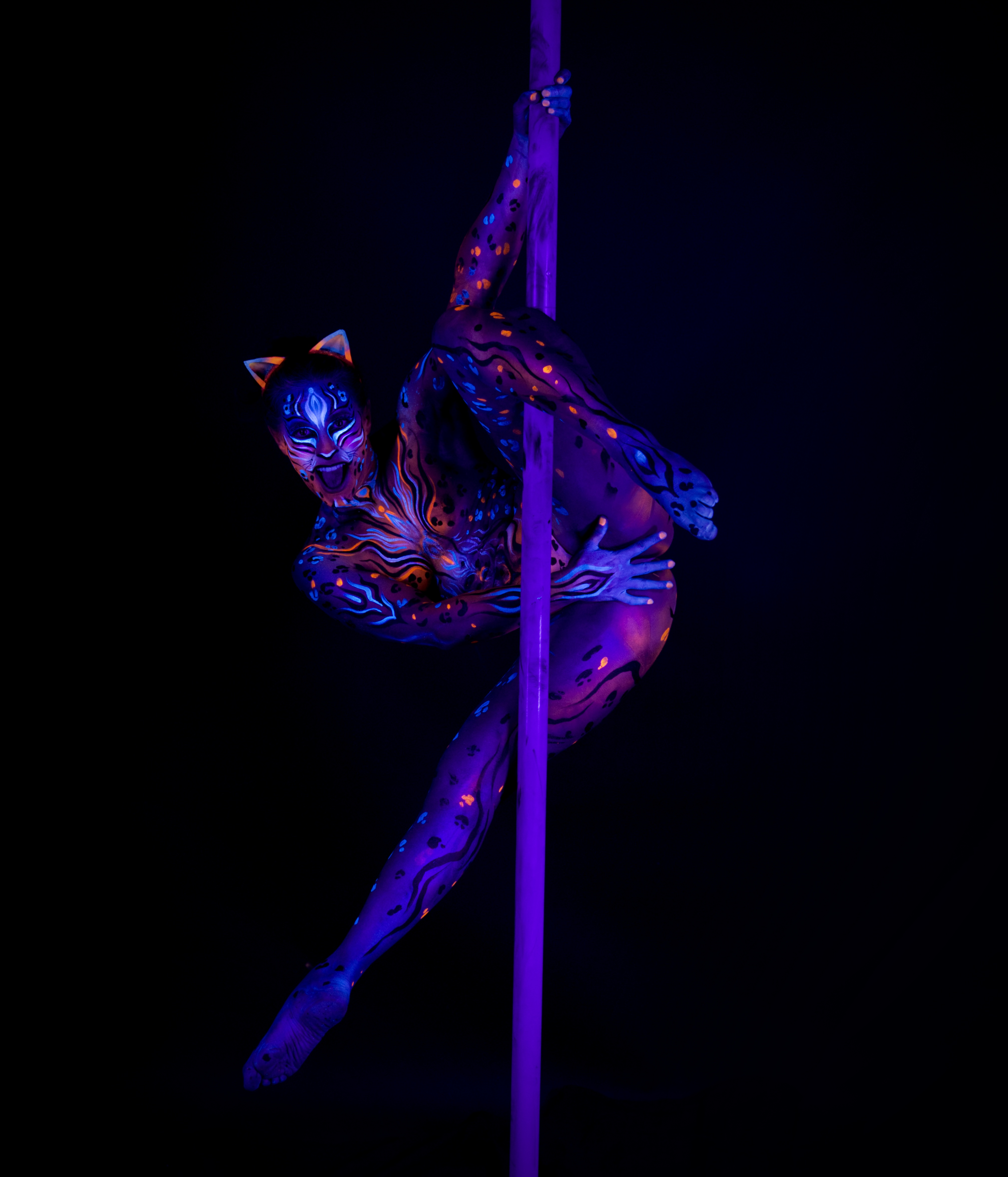 Marcela is curled against a pole in a crouched acrobatic pose with her knees drawn in, wearing cat-ear prosthetics. Her body is painted with a blue and orange leopard-spot pattern. Bodypaint by Joana Rohr, Julia. Photographed by Elad Itzkin Photography under UV blacklight in his studio in Gesundbrunnen, Berlin.