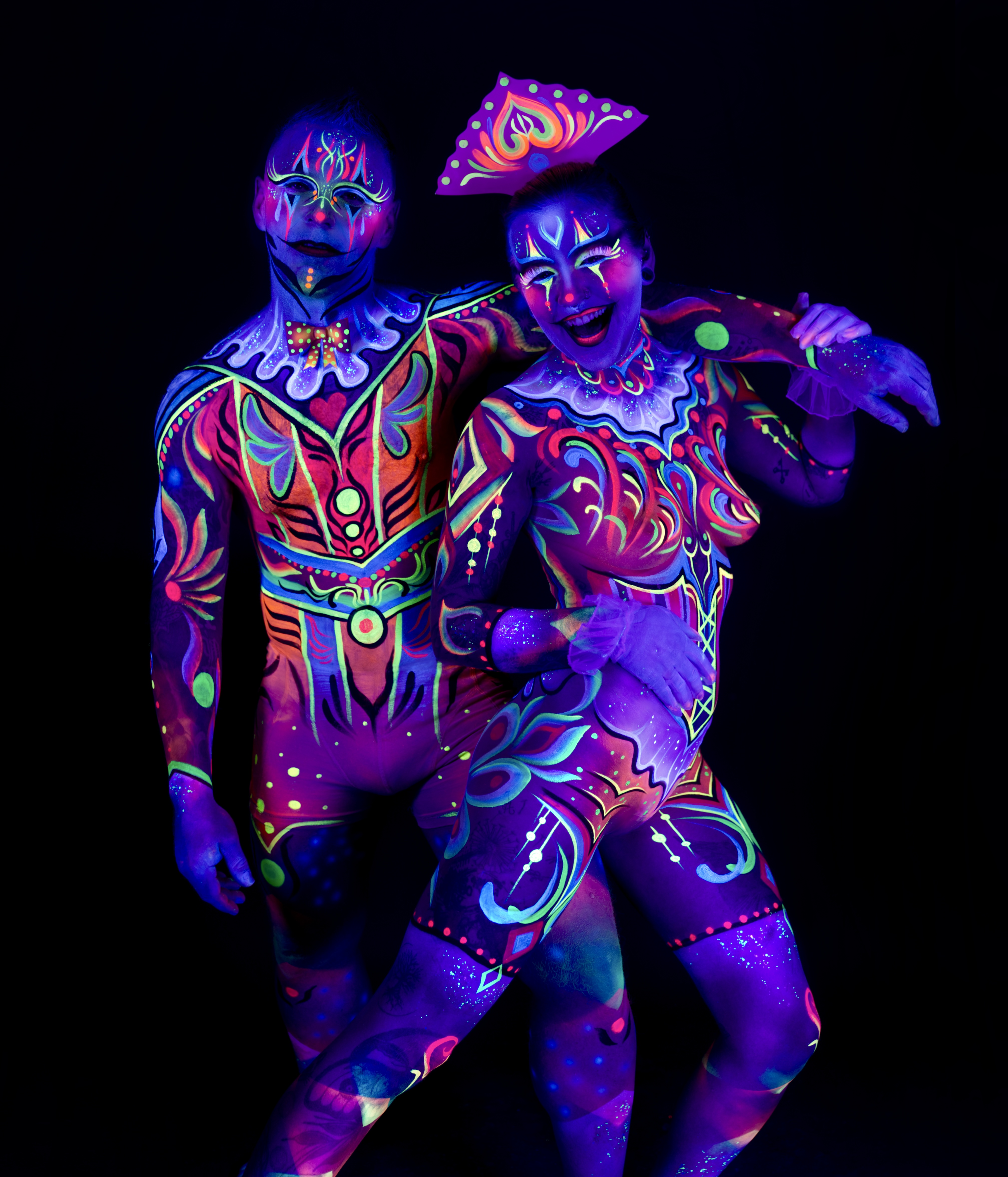 Laura and Francis stand together in an upright pose. Their bodies are painted in an ornate harlequin-style design with orange, purple, and green geometric and floral patterns, and Laura wears a folded headpiece. Bodypaint by Joana Rohr, Sage Brewer. Photographed by Elad Itzkin Photography under UV blacklight in his studio in Gesundbrunnen, Berlin.