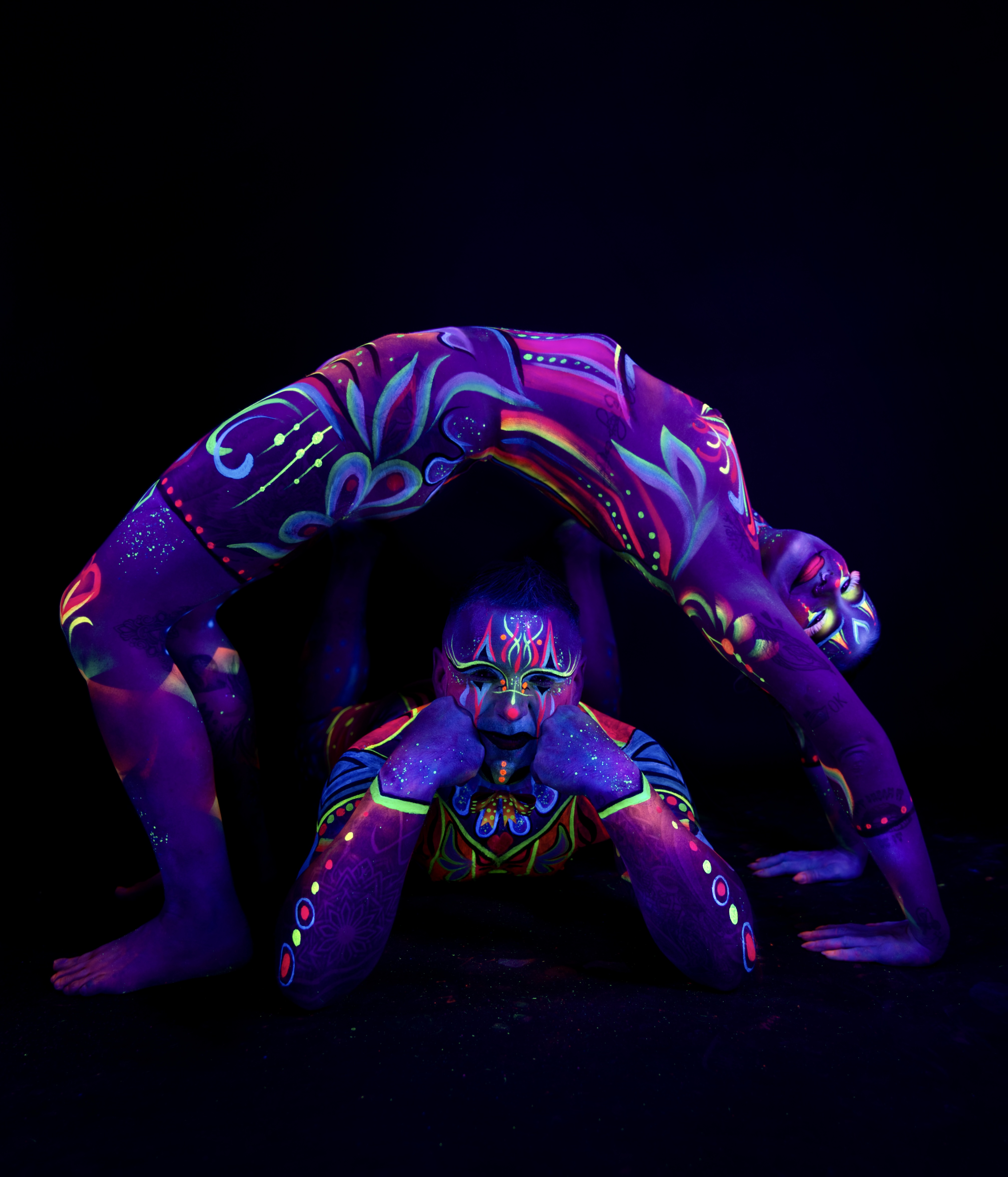 Laura and Francis pose together, one arched backward in a bridge position forming an arch over the other, who kneels beneath. Both are painted in a purple design with colorful floral and geometric patterns. Bodypaint by Joana Rohr, Sage Brewer. Photographed by Elad Itzkin Photography under UV blacklight in his studio in Gesundbrunnen, Berlin.