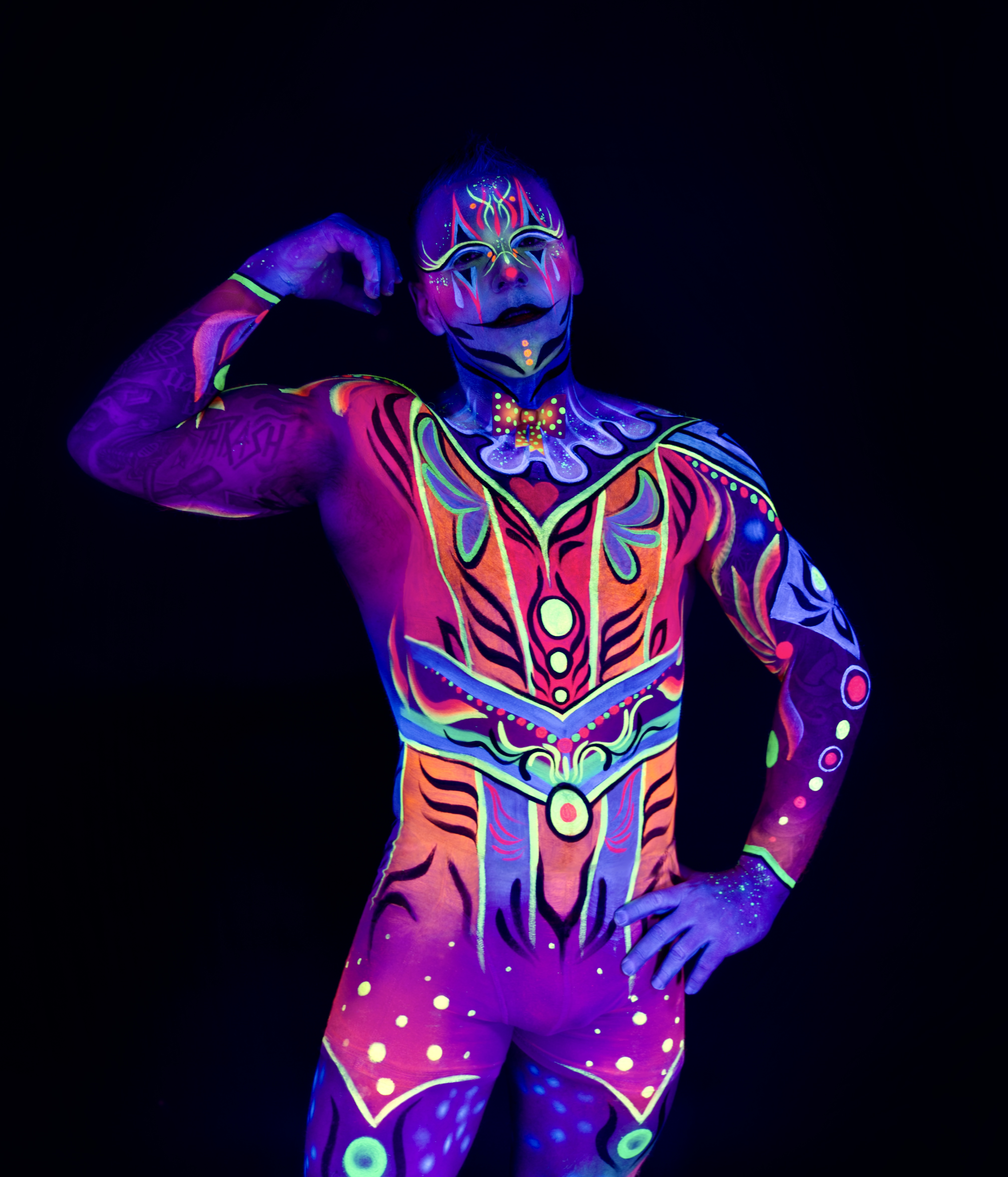 Francis stands in a three-quarter pose against a black background. The bodypaint forms a symmetrical, mask-like pattern across the face and torso in orange, purple and blue tones, suggesting a stylized tribal design. Bodypaint by Joana Rohr and Sage Brewer. Photographed by Elad Itzkin Photography under UV light in his studio in Gesundbrunnen, Berlin.