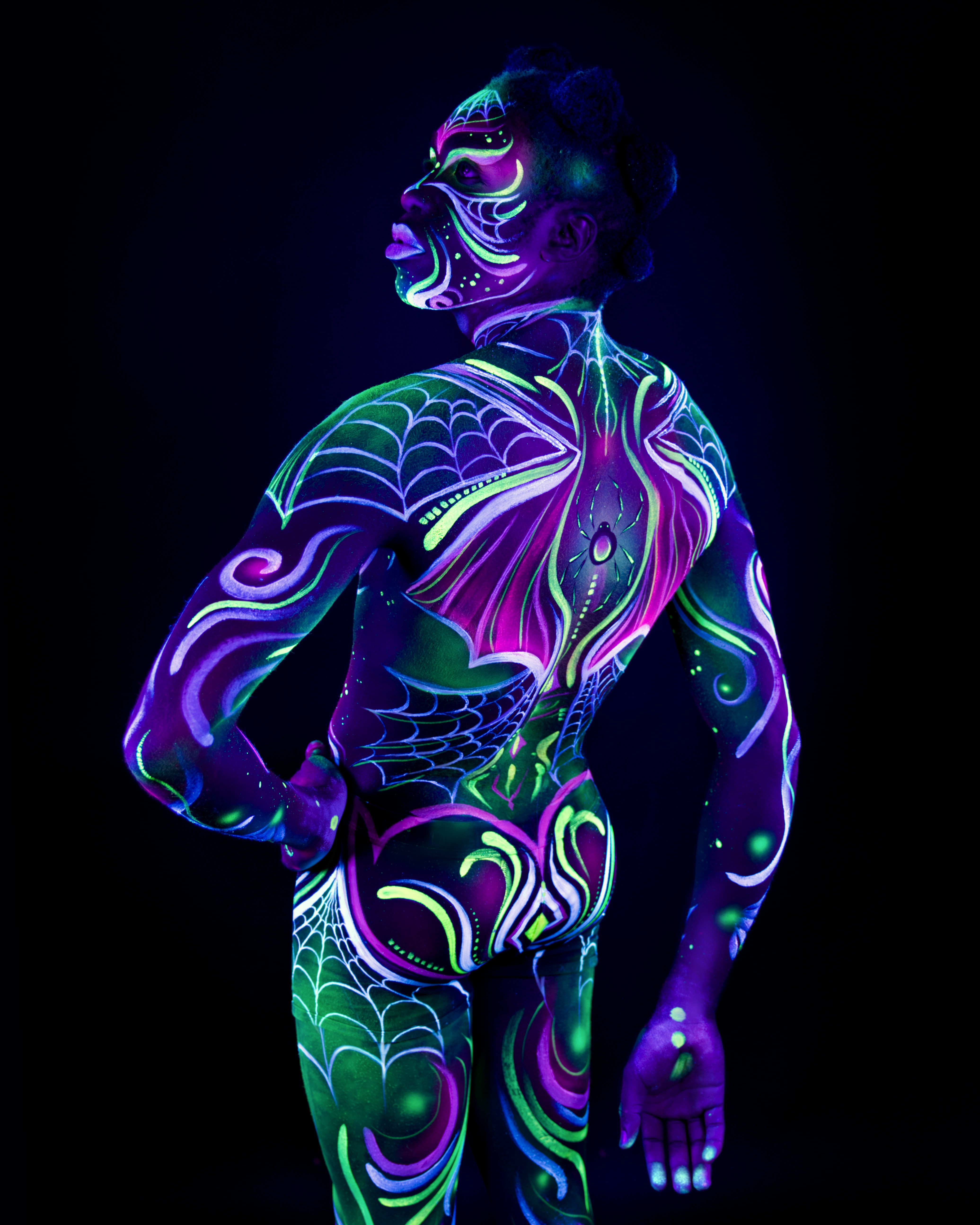 Bikme stands with the back to the camera, head turned to the side in profile. The bodypaint covers the back and legs in a purple and green design combining a spiderweb pattern with a wing-shaped motif across the shoulders. Bodypaint by Joana Rohr and Sage Brewer. Photographed by Elad Itzkin Photography under UV light in his studio in Gesundbrunnen, Berlin.