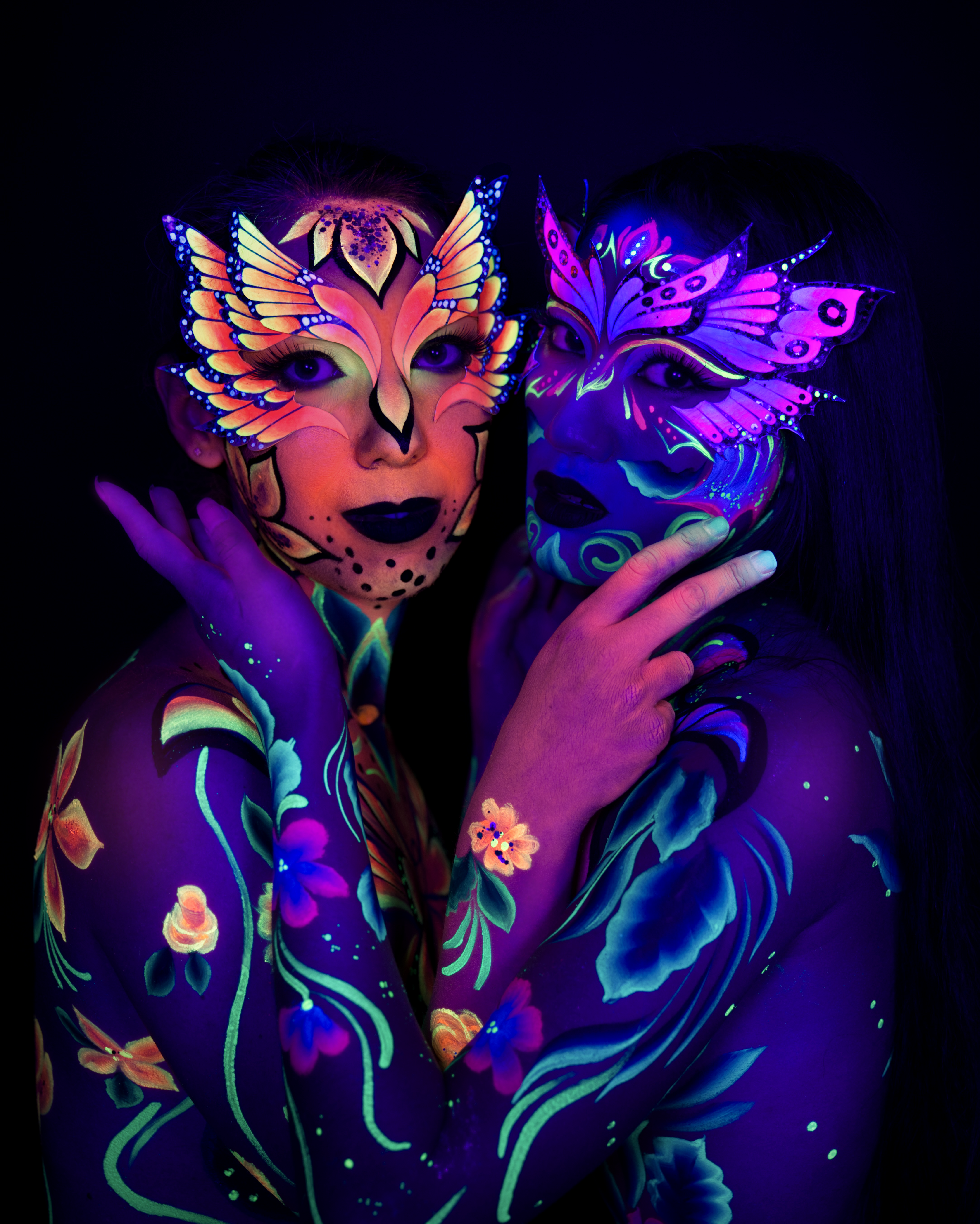 Alessia and Daryna stand close together facing the camera, cropped at the upper body. Each wears a butterfly-wing face design, one rendered in orange and yellow and the other in pink and purple, paired with floral vine bodypaint in green, purple and teal. Bodypaint by Joana Rohr and Sage Brewer. Photographed by Elad Itzkin Photography under UV light in his studio in Gesundbrunnen, Berlin.