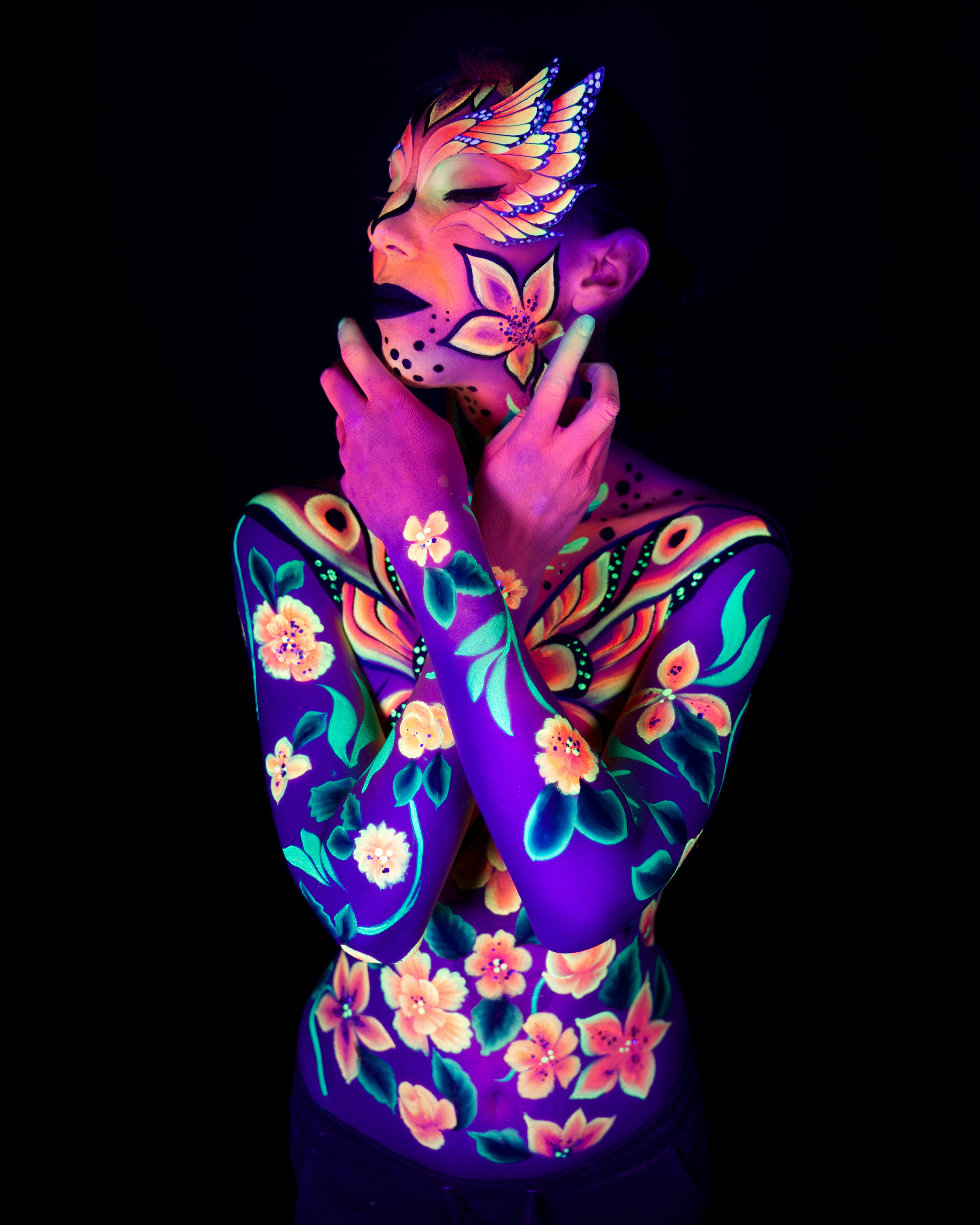 Alessia is photographed facing the camera in an upper-body crop. An orange and yellow wing-shaped design frames the face, extending into a purple bodypaint pattern with orange and green floral accents across the shoulders and chest. Bodypaint by Joana Rohr and Sage Brewer. Photographed by Elad Itzkin Photography under UV light in his studio in Gesundbrunnen, Berlin.