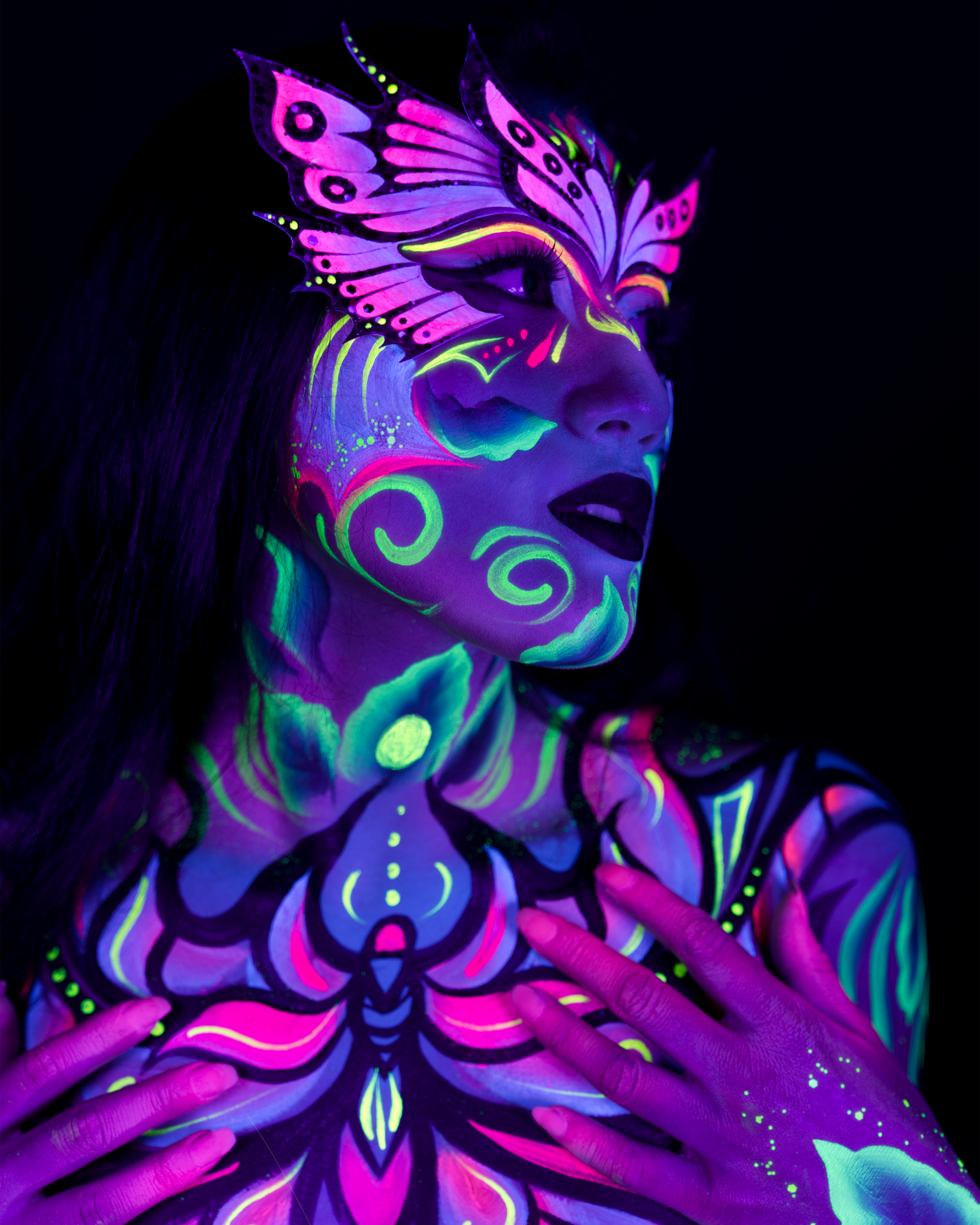 Daryna is shown in a close upper-body crop, facing the camera at a slight angle. The design includes a pink and purple butterfly-wing motif on the face and a floral pattern in pink, purple and teal across the chest and neck. Bodypaint by Joana Rohr and Sage Brewer. Photographed by Elad Itzkin Photography under UV light in his studio in Gesundbrunnen, Berlin.