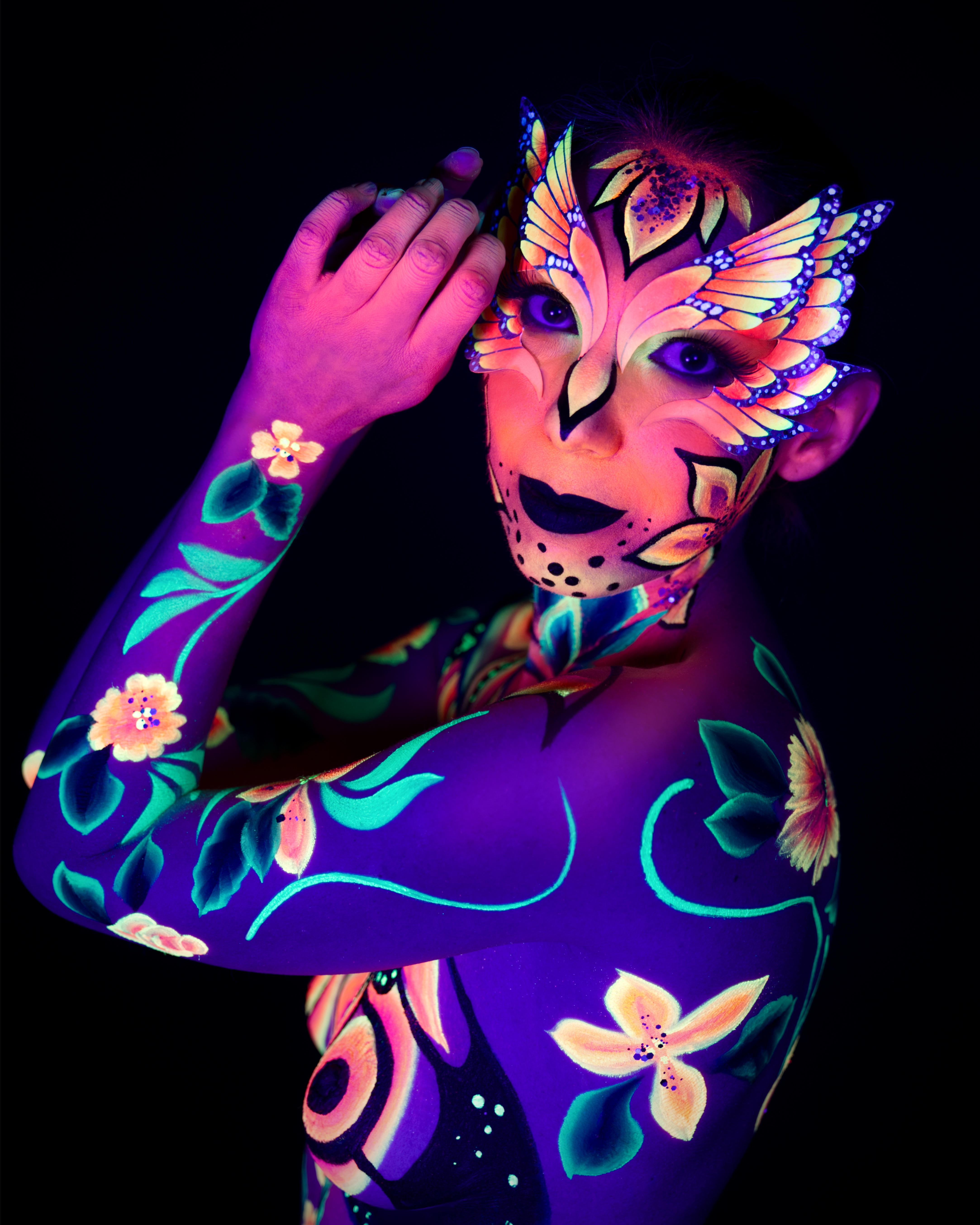Alessia is photographed in a three-quarter turn, cropped at the upper body. An orange and yellow wing-shaped face design continues into a floral bodypaint pattern in purple, teal and orange across the shoulder and chest. Bodypaint by Joana Rohr and Sage Brewer. Photographed by Elad Itzkin Photography under UV light in his studio in Gesundbrunnen, Berlin.